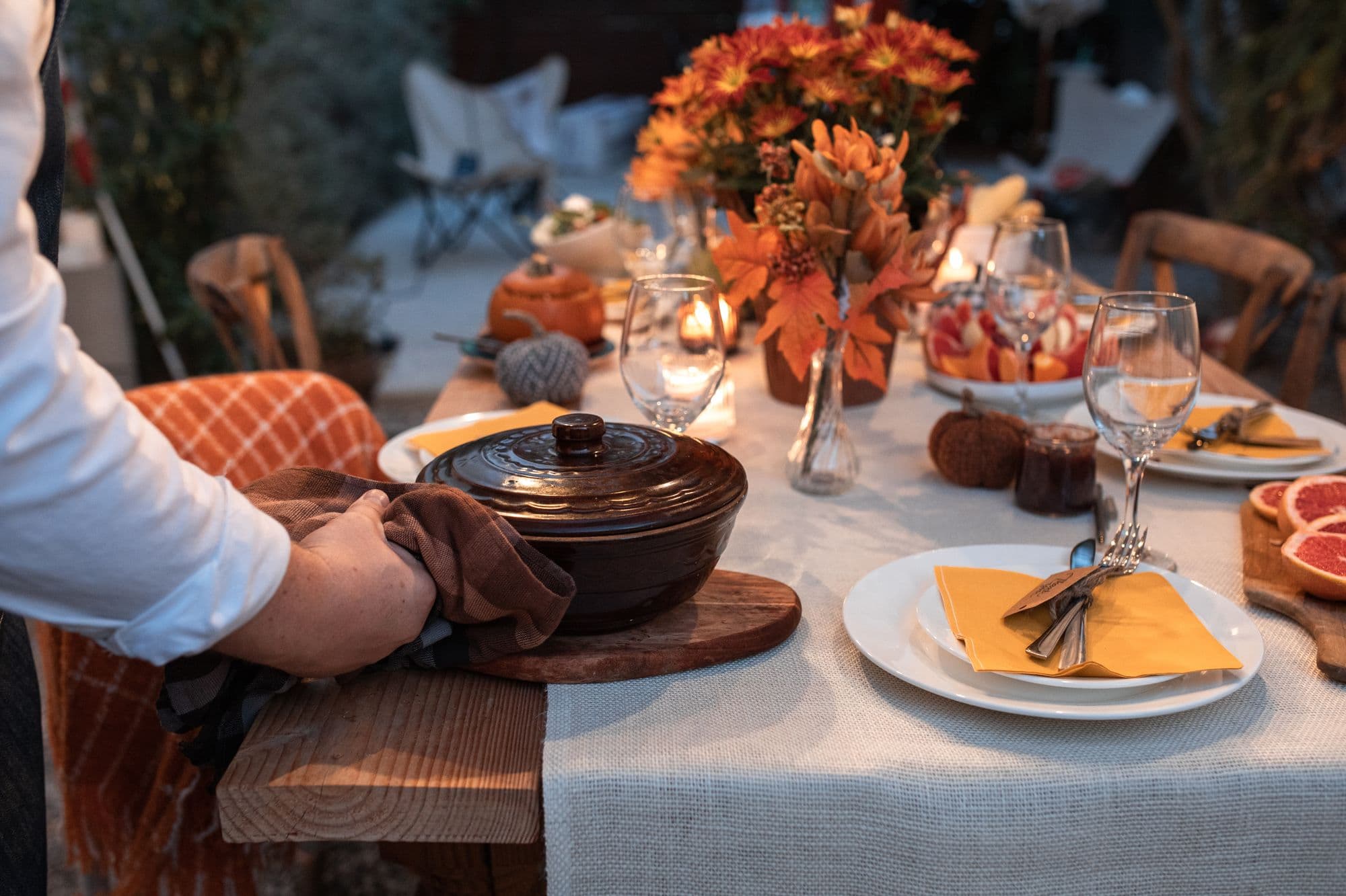 Outdoor autumn dinner table with orange flowers, candles and plates as a host sets down a covered pot