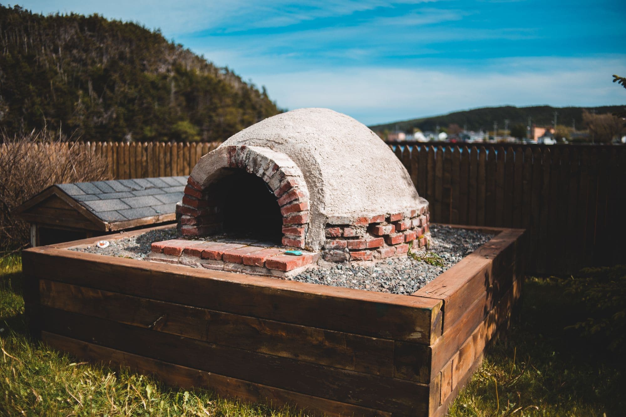 Homemade brick-and-mortar dome oven on a gravel bed inside a timber-framed raised base in a backyard