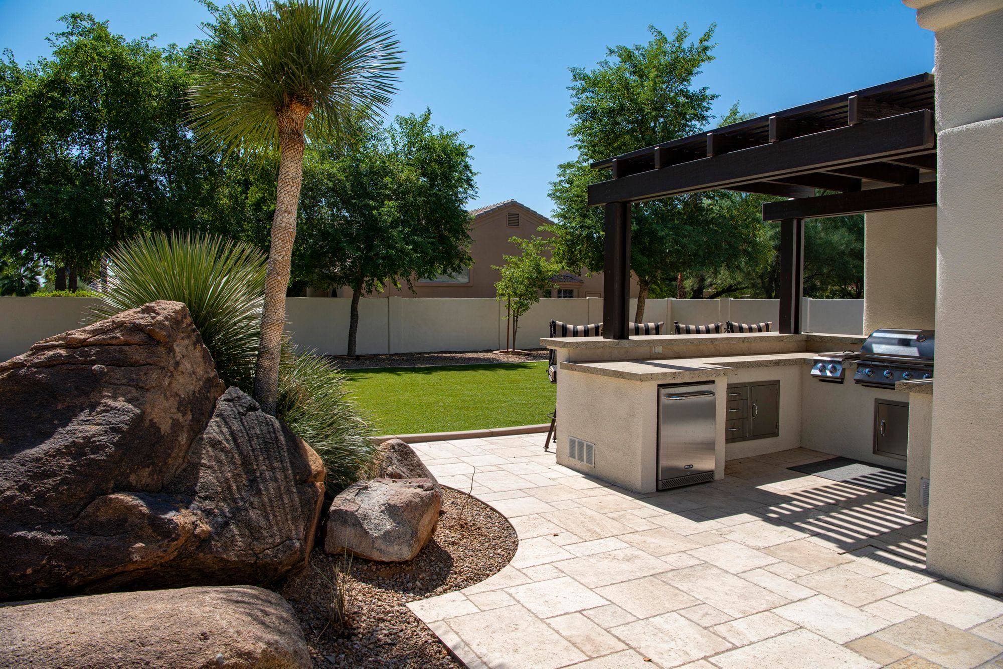 L-shaped stucco outdoor kitchen with a raised bar ledge, a built-in gas grill, a double side burner, drawers, access doors and an under-counter fridge beside a dark pergola on a travertine patio