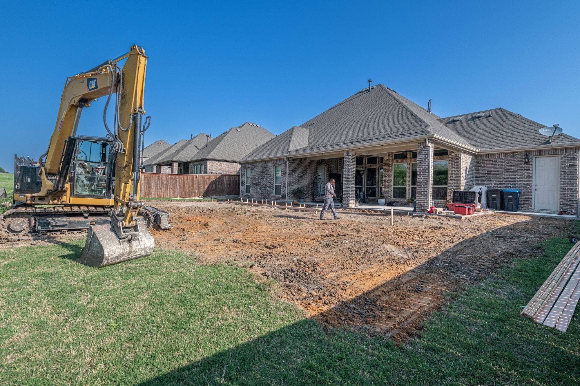 Excavator grading a cleared backyard behind a brick house in preparation for a new patio