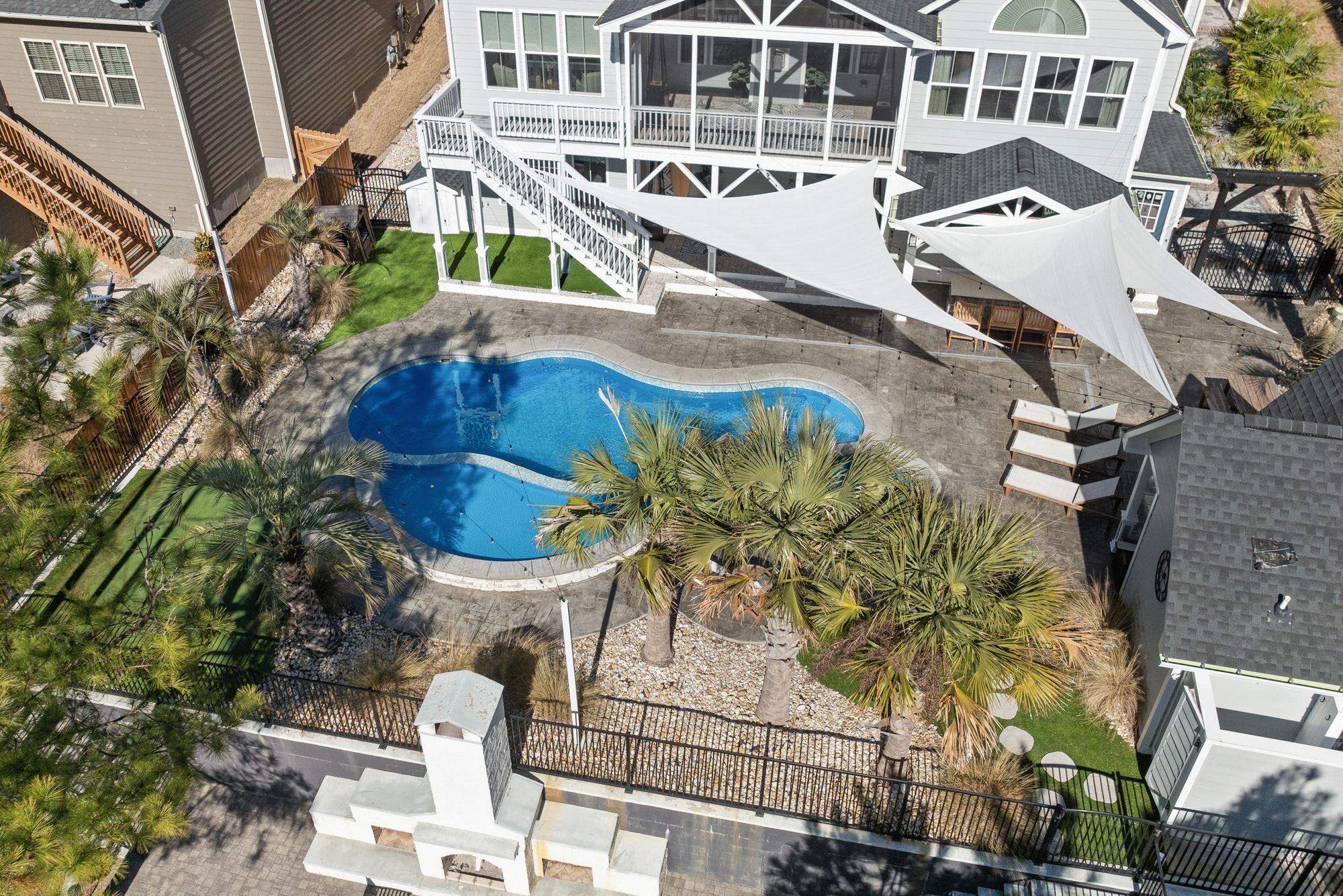 Aerial view of a backyard with a kidney-shaped pool, palm plantings, shade sails and a white masonry outdoor fireplace with side counters in the corner