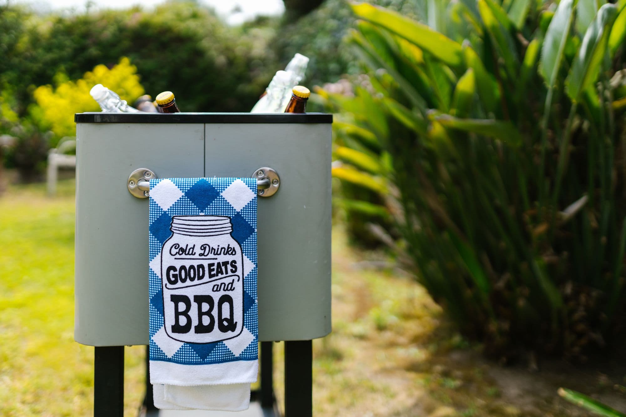 Grey stand-up beverage cooler filled with bottles and ice, with a "Good Eats and BBQ" towel hanging on the front, in a garden