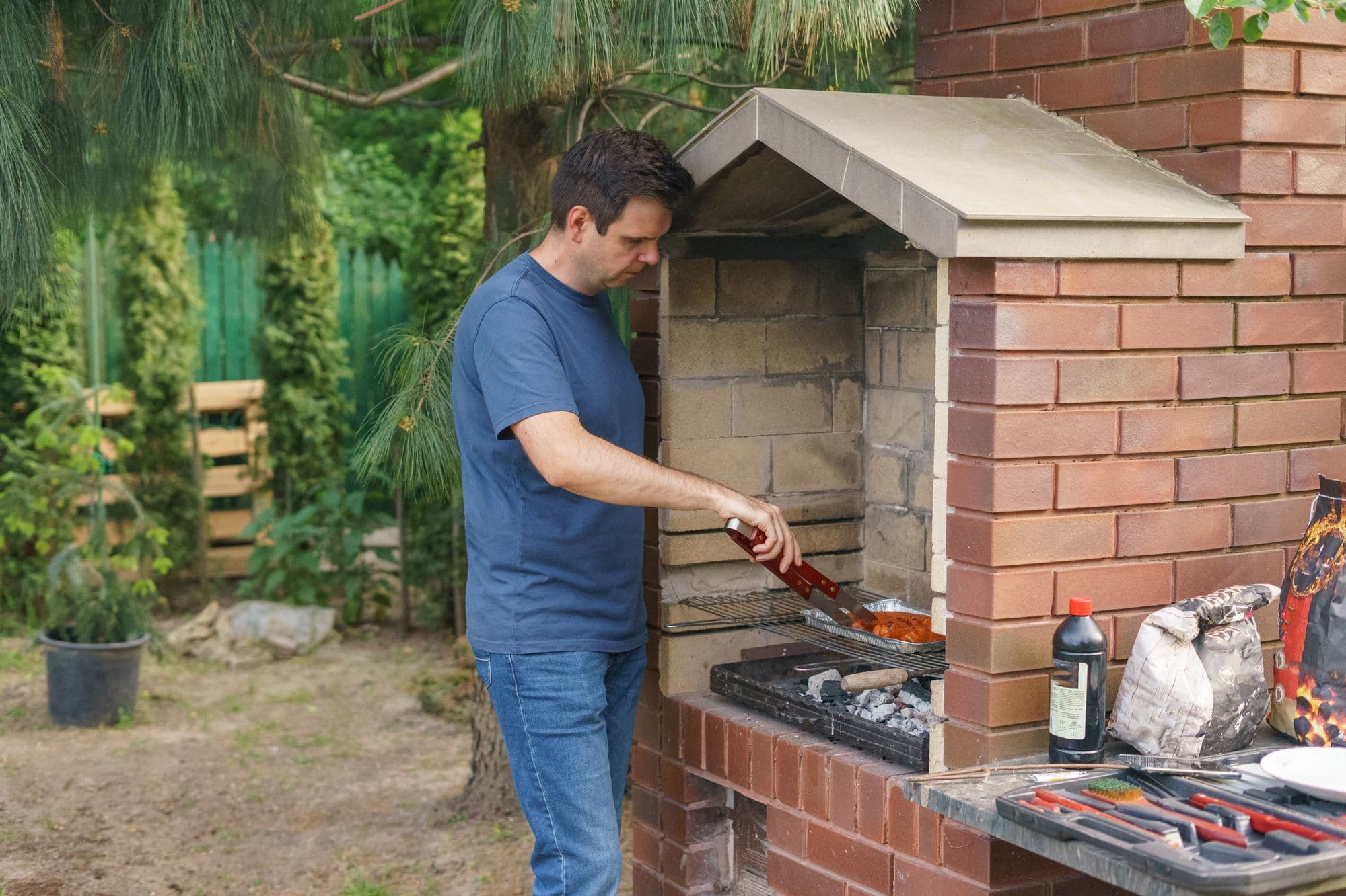 Man cooking on a charcoal grill grate inside a built brick barbecue with a stone hood in a backyard garden