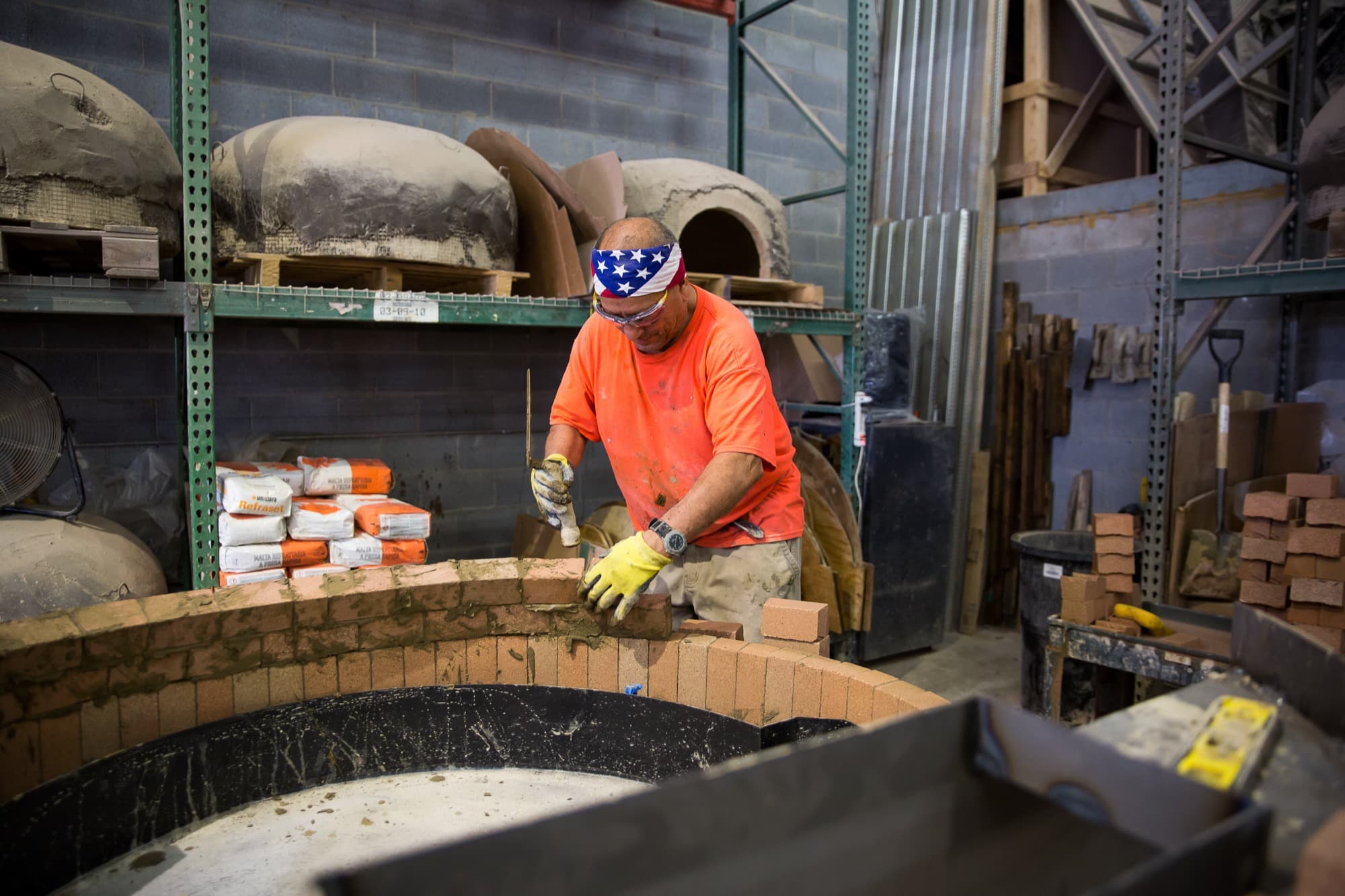 Mason in an orange shirt laying the firebrick ring of a wood-fired oven in a workshop, with finished oven domes on shelves behind