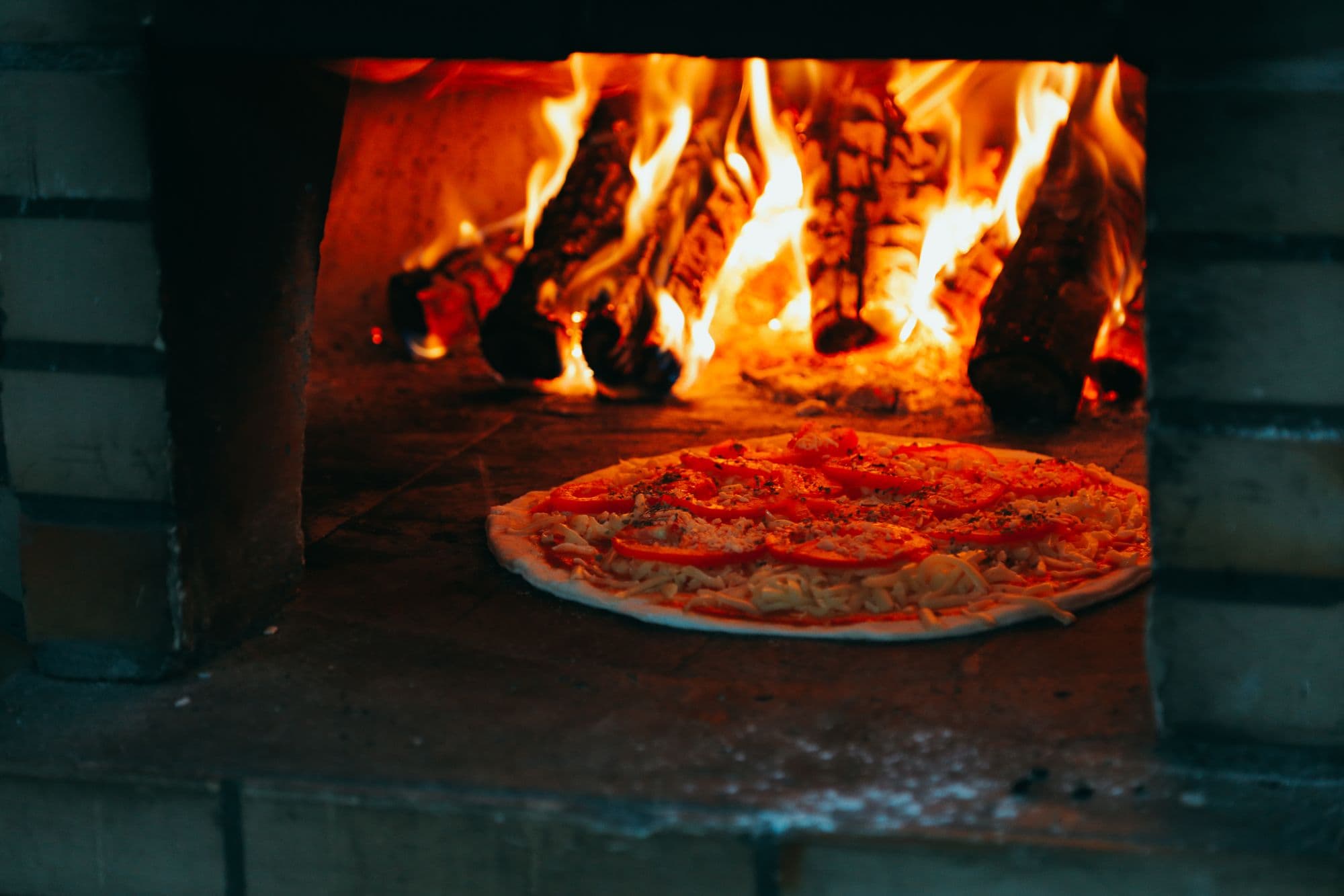 Tomato pizza on the floor of a brick oven in front of a blazing wood fire