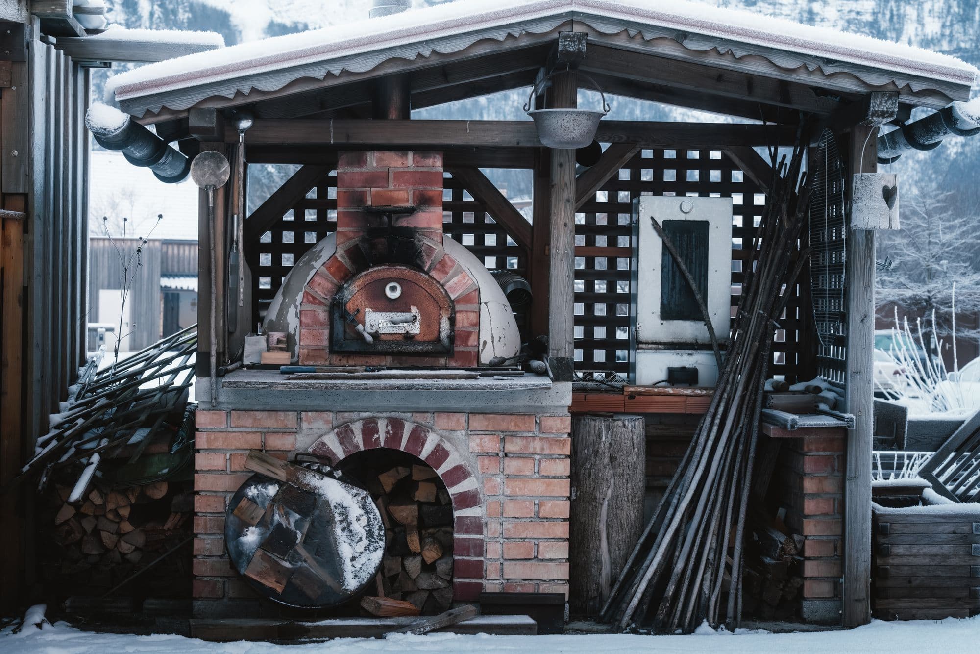 Brick wood-fired oven and firewood store under a wooden shelter, with snow on the roof and ground