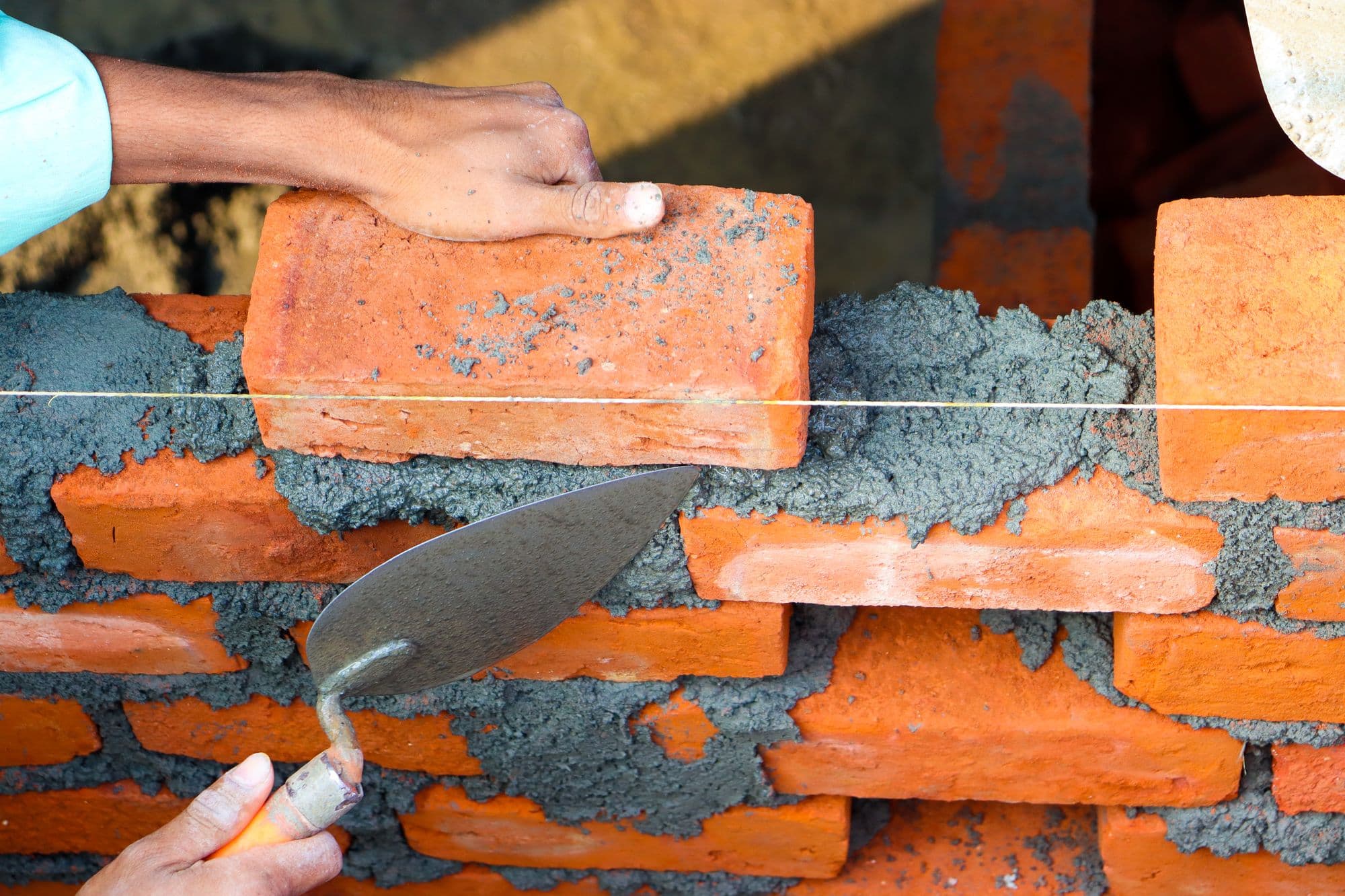 Hands setting a red brick into fresh mortar along a string line with a trowel