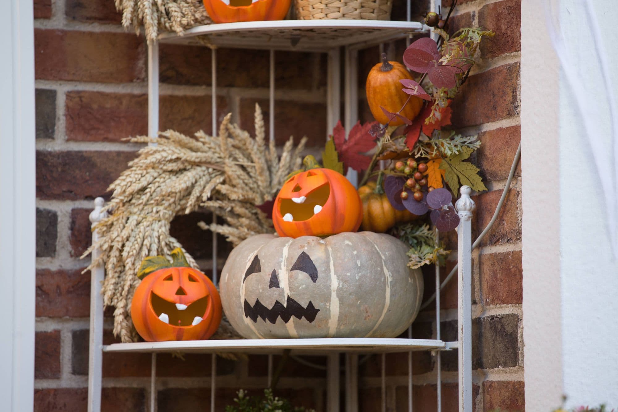 Carved and painted pumpkins with wheat and autumn leaves on a white shelf against a brick wall