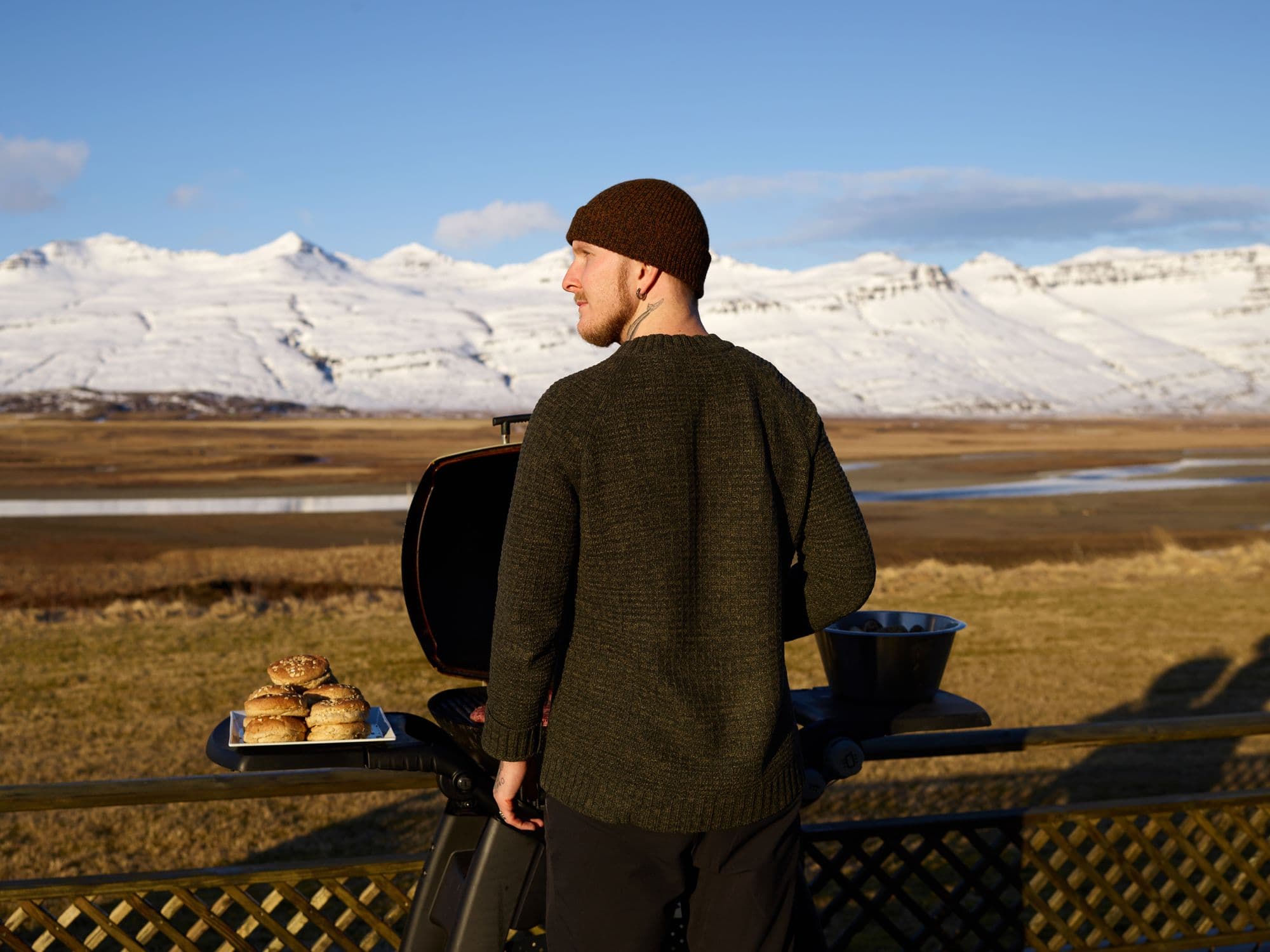 Man in a knit cap at a grill on a deck with a tray of buns, overlooking snow-capped mountains