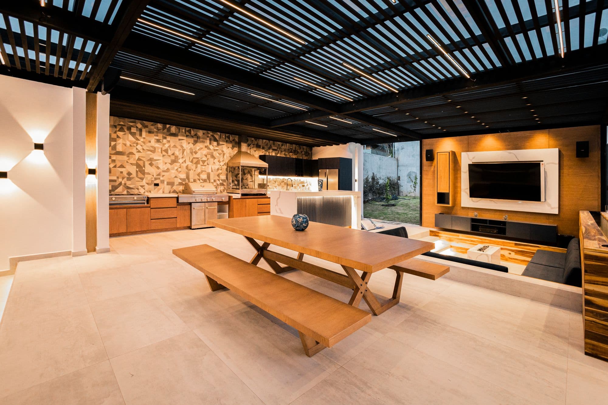 Covered outdoor kitchen under a louvered roof with wood-front base cabinets, a stainless built-in grill, a geometric tile backsplash and a long wooden dining table with benches