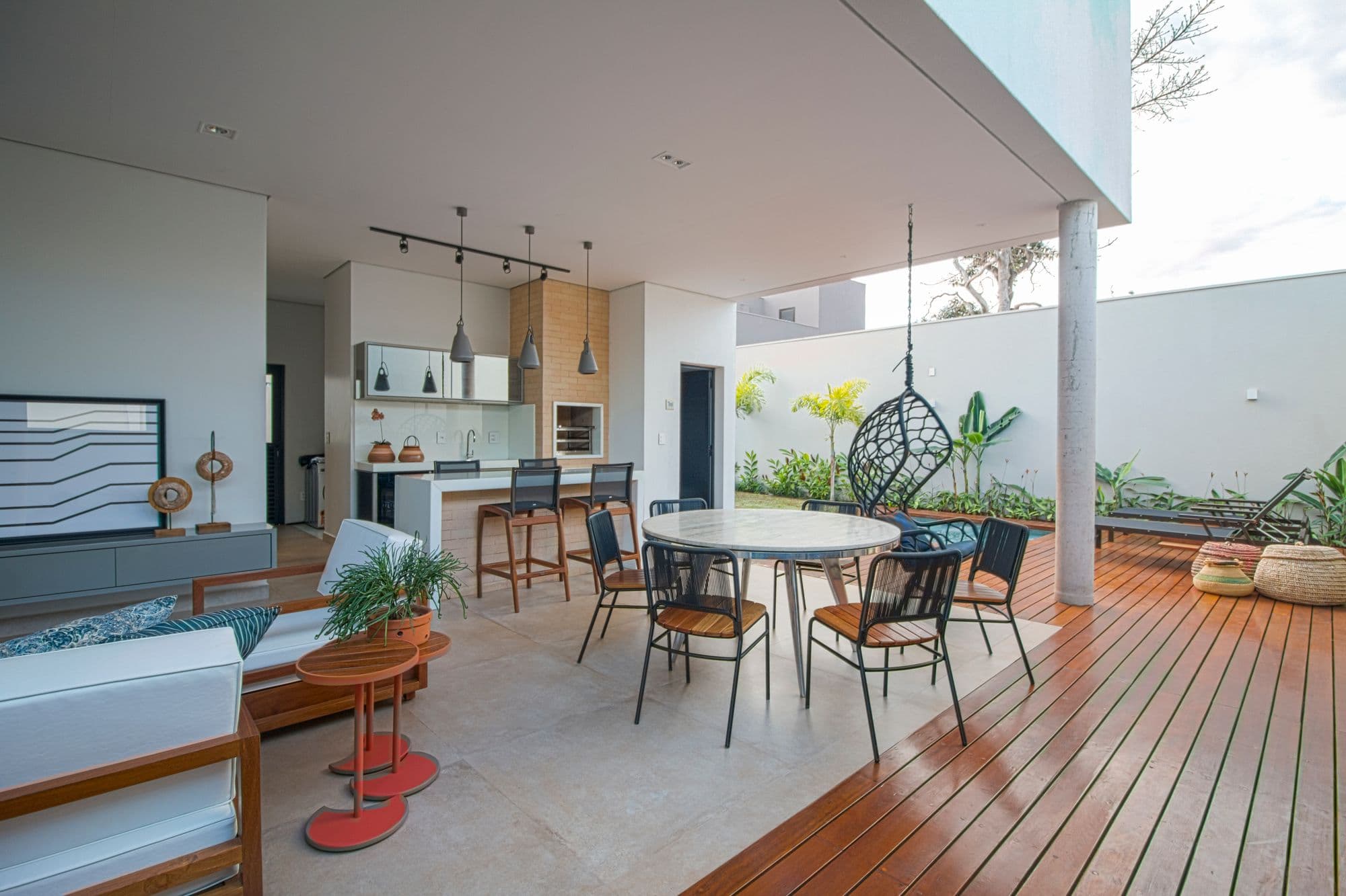 Contemporary covered patio with an open kitchen counter and bar stools, a round dining table with chairs, a hanging egg chair and a wooden deck