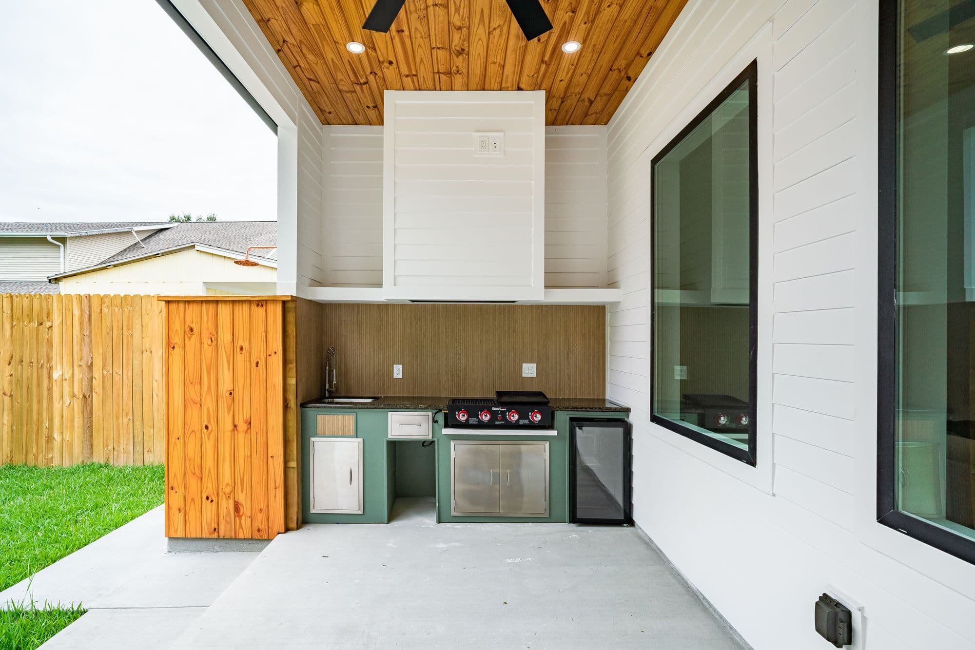 Small covered-patio outdoor kitchen with green base cabinets, a built-in flat-top griddle, stainless access doors and a glass-door beverage fridge beneath a wood-plank ceiling