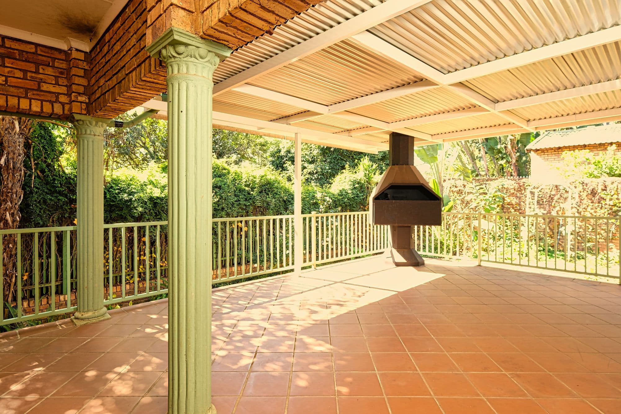 Covered tiled patio with a corrugated roof, classical columns, iron railings and a freestanding hooded masonry barbecue