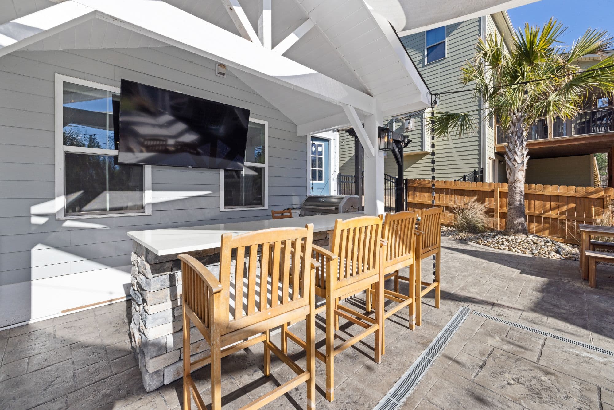 Covered patio with a stacked-stone bar island, a light quartz bar top, wooden bar stools and a wall-mounted TV, with a stainless gas grill at the far end