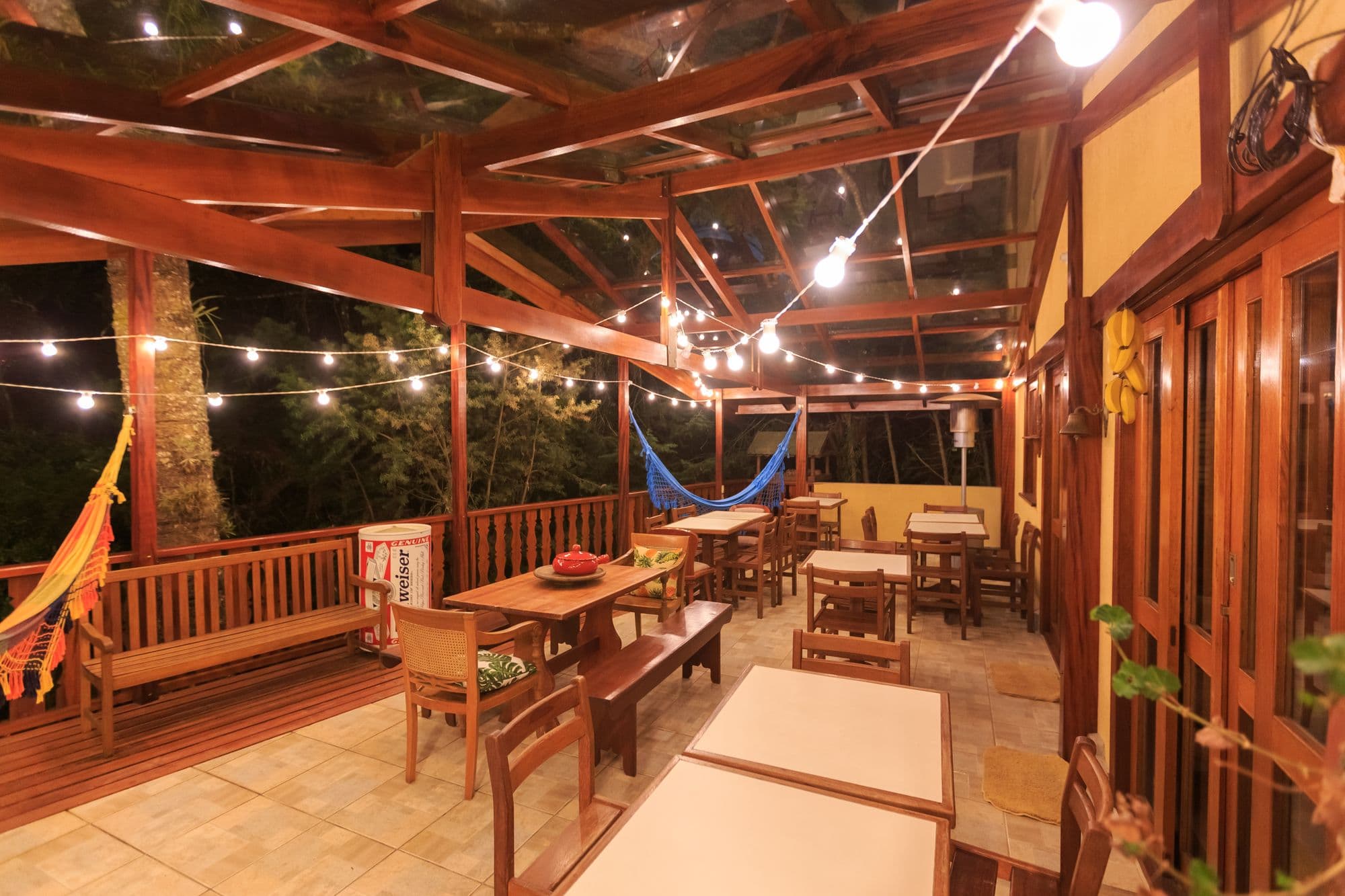 Covered wooden patio at night with globe string lights, dining tables, benches and hammocks