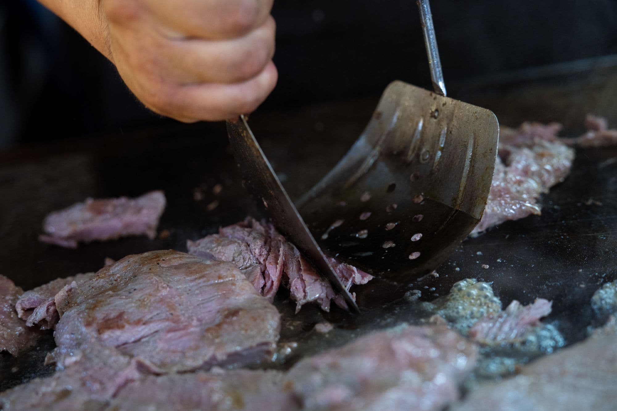 Hand using a metal spatula and scraper to turn thin beef slices on a dark flat-top griddle