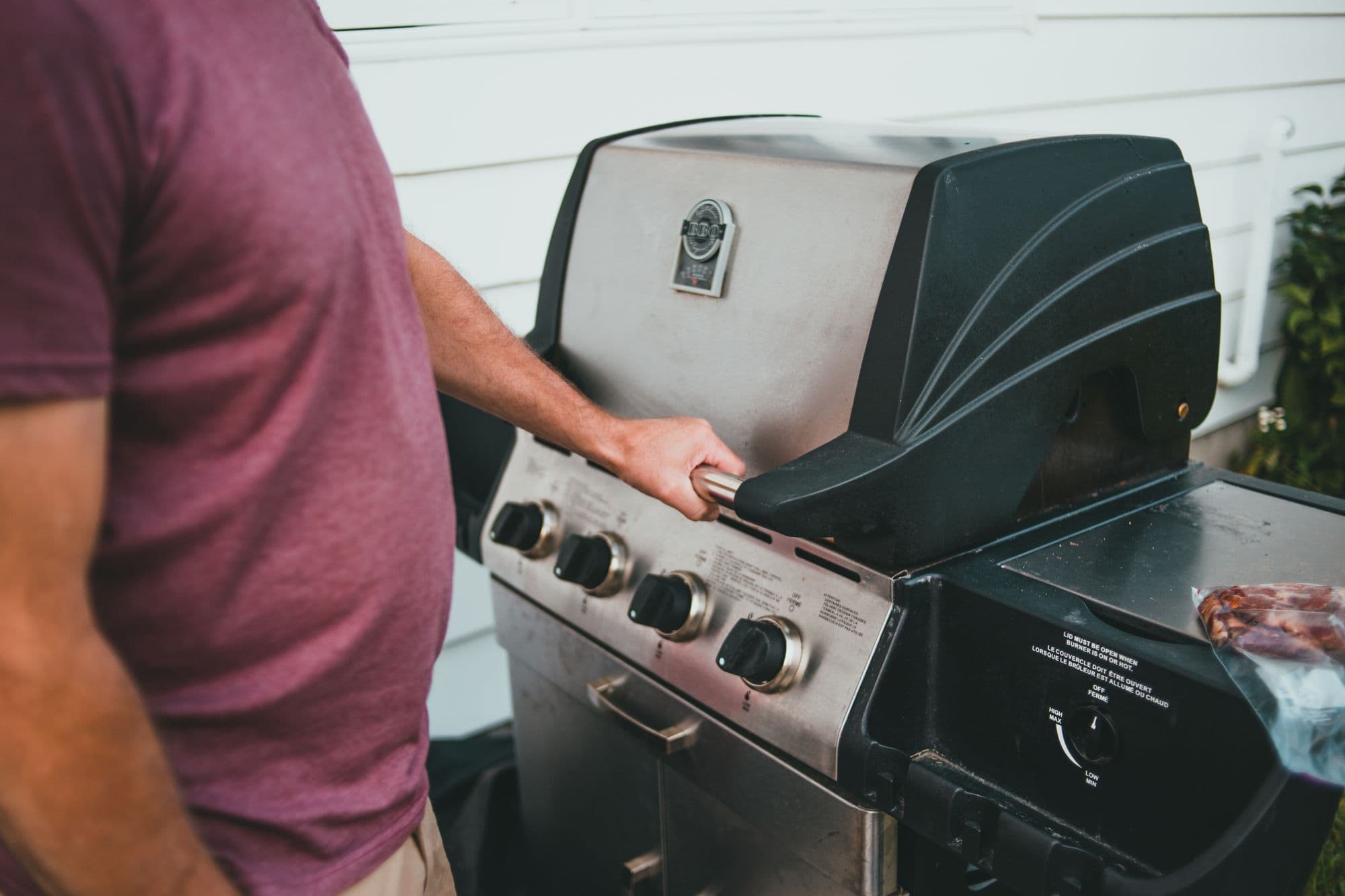 Person standing at a stainless steel and black freestanding four-burner gas grill beside white siding