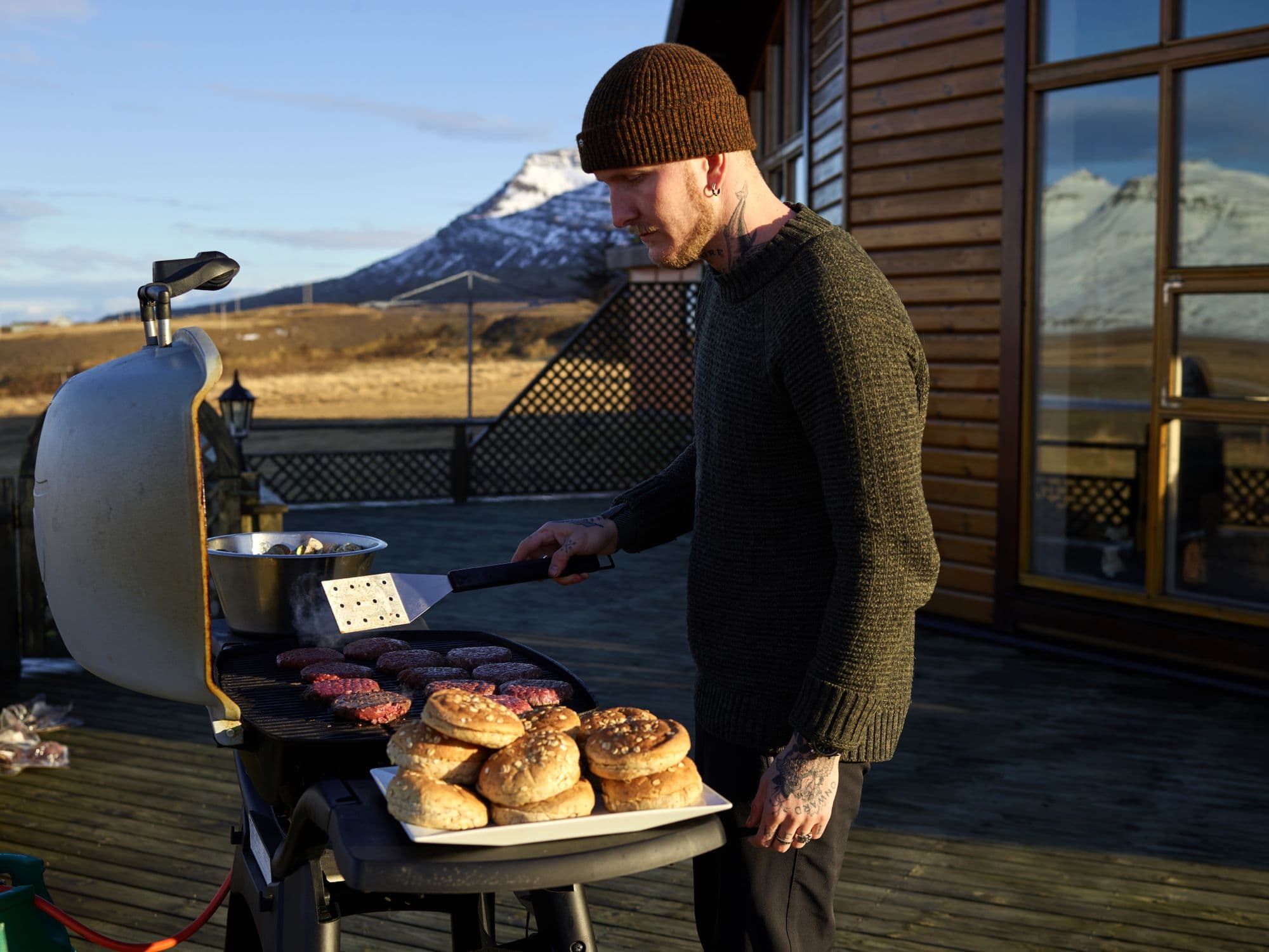 Man in a knit cap cooking burgers on a flat-top griddle on a wooden deck, with a tray of buns and snow-capped mountains behind