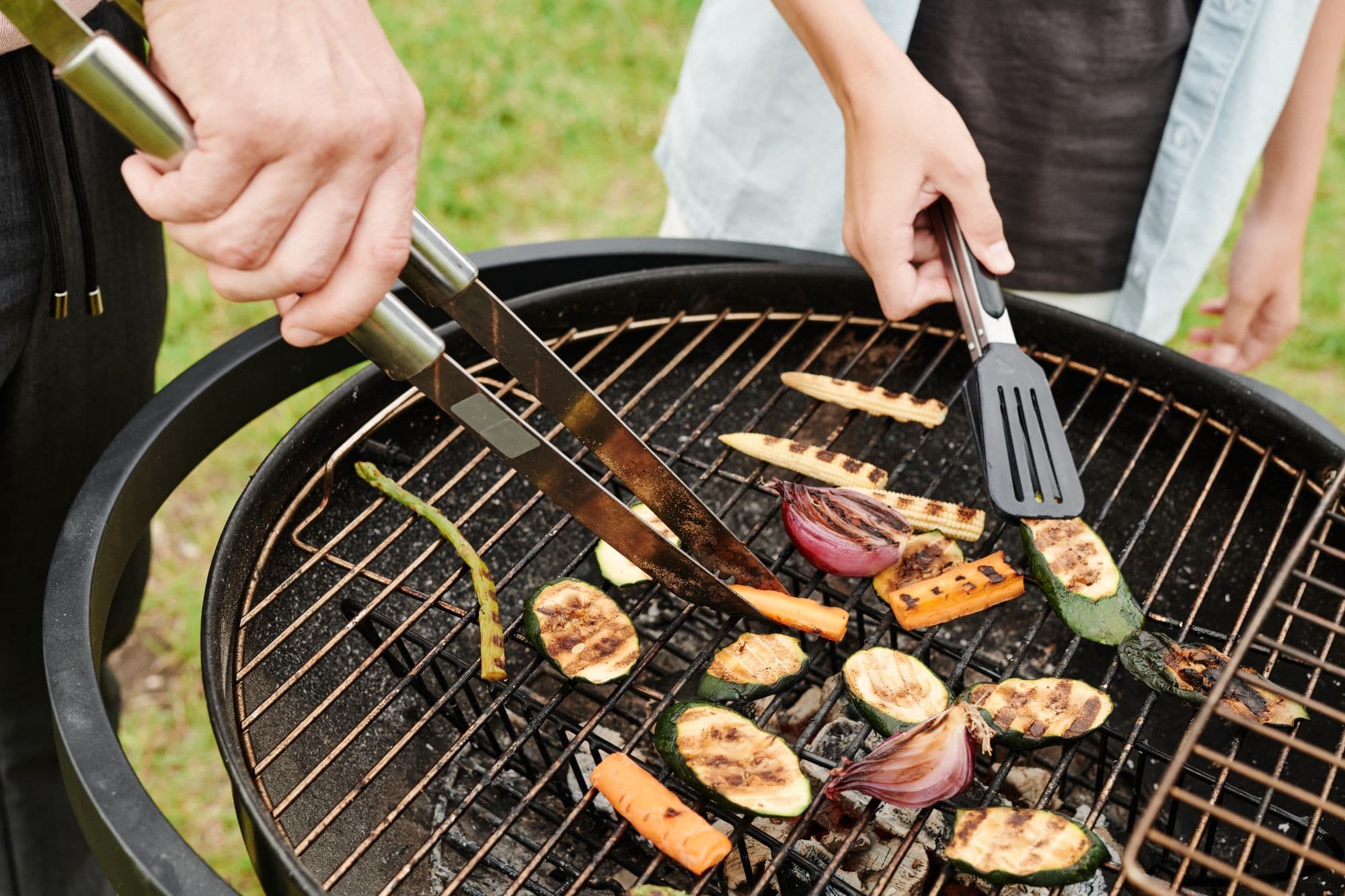 Hands with tongs and a spatula turning zucchini, carrots and onion on a round charcoal grill