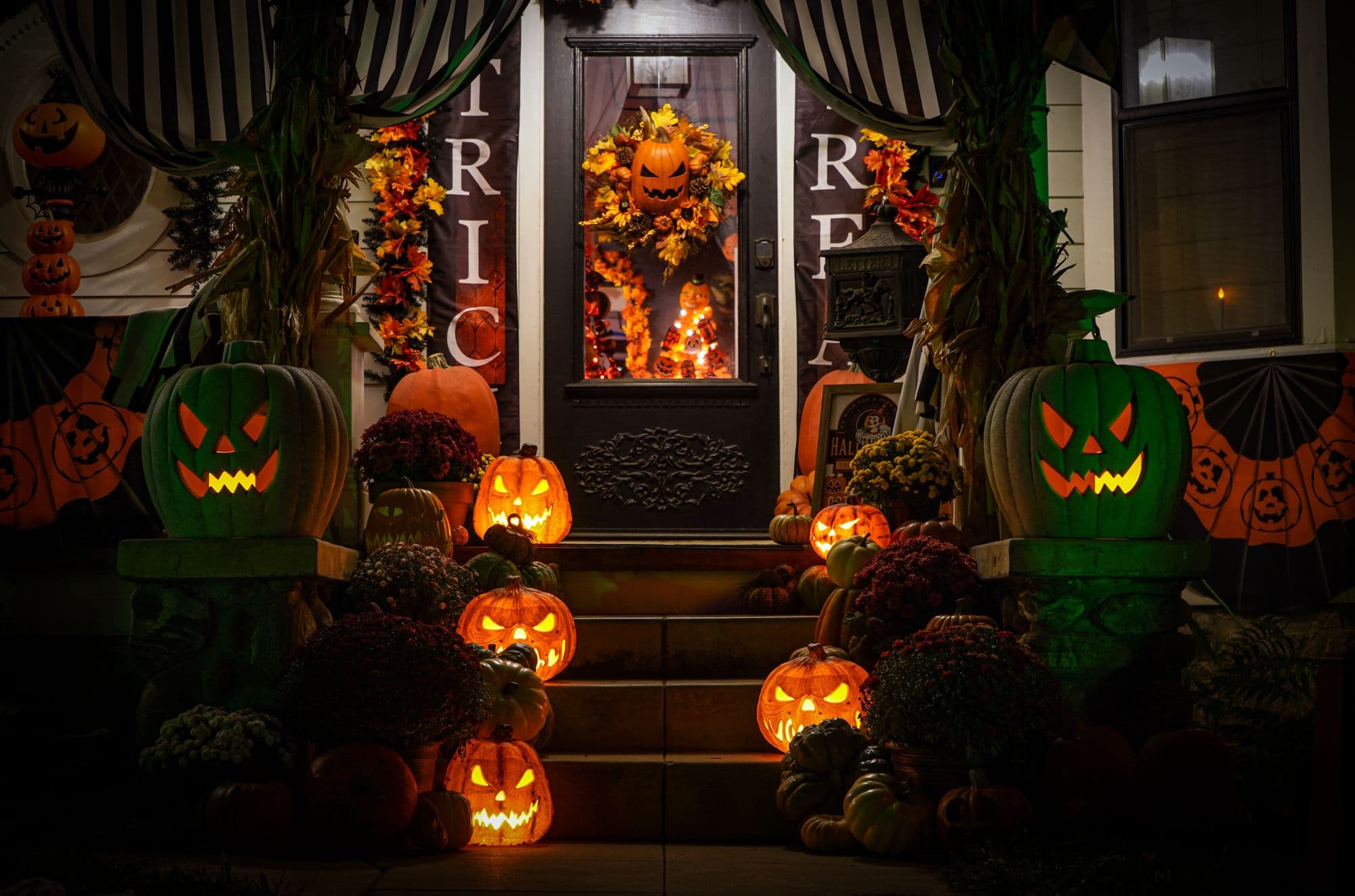 Porch at night with glowing carved jack-o'-lanterns, mums and a Halloween-decorated front door
