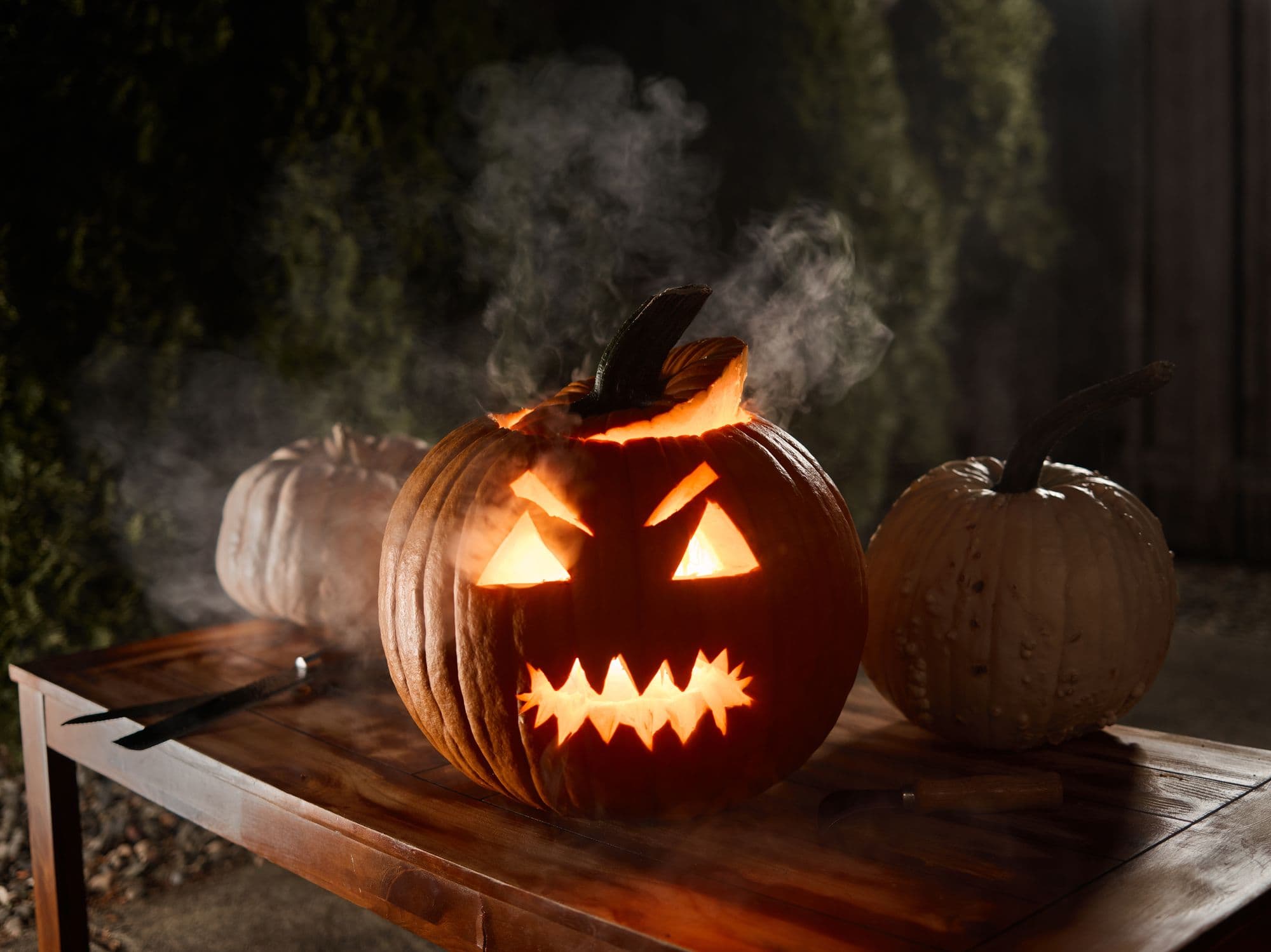 Glowing carved jack-o'-lantern with smoke rising, between two pumpkins on an outdoor wooden table at night