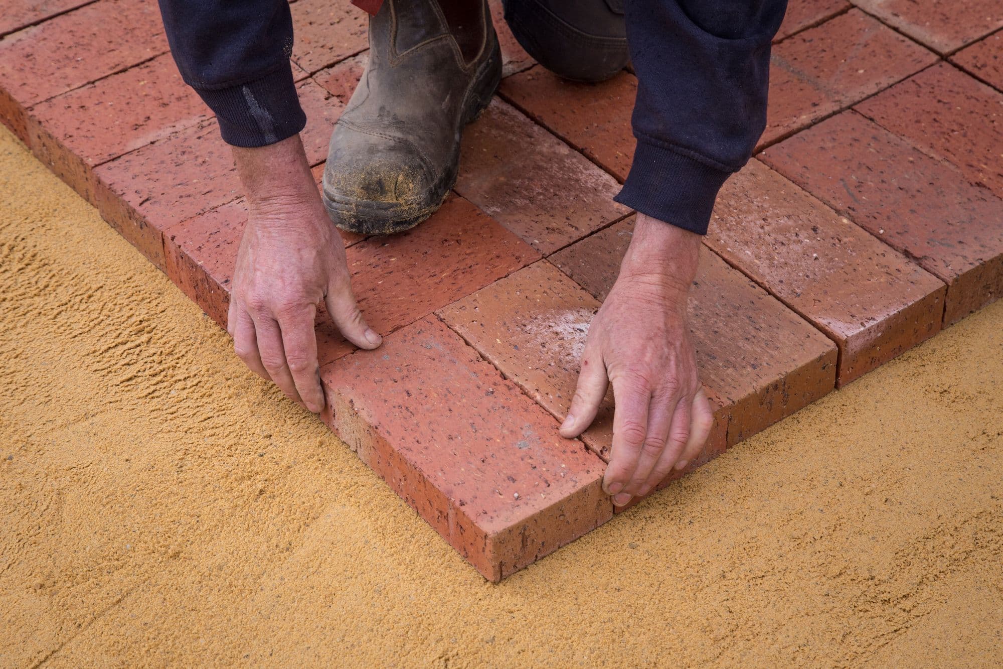 Gloved worker's hands setting red clay brick pavers on a sand bed