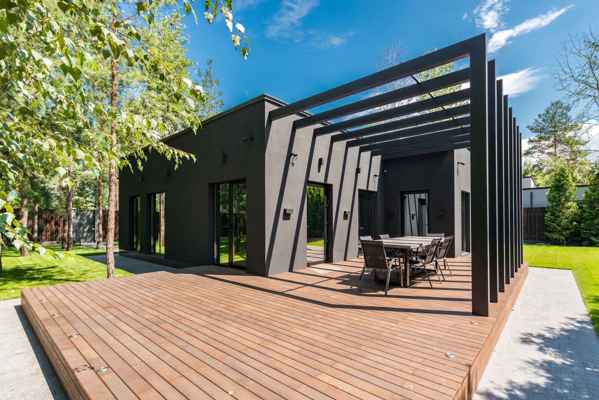Modern dark house with a wide wooden deck, a slatted pergola and an outdoor dining set beside the lawn