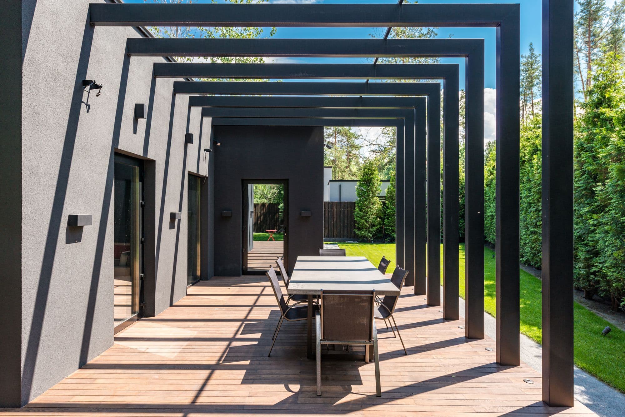 Modern black steel pergola over a wooden deck with an outdoor dining table and chairs beside a lawn