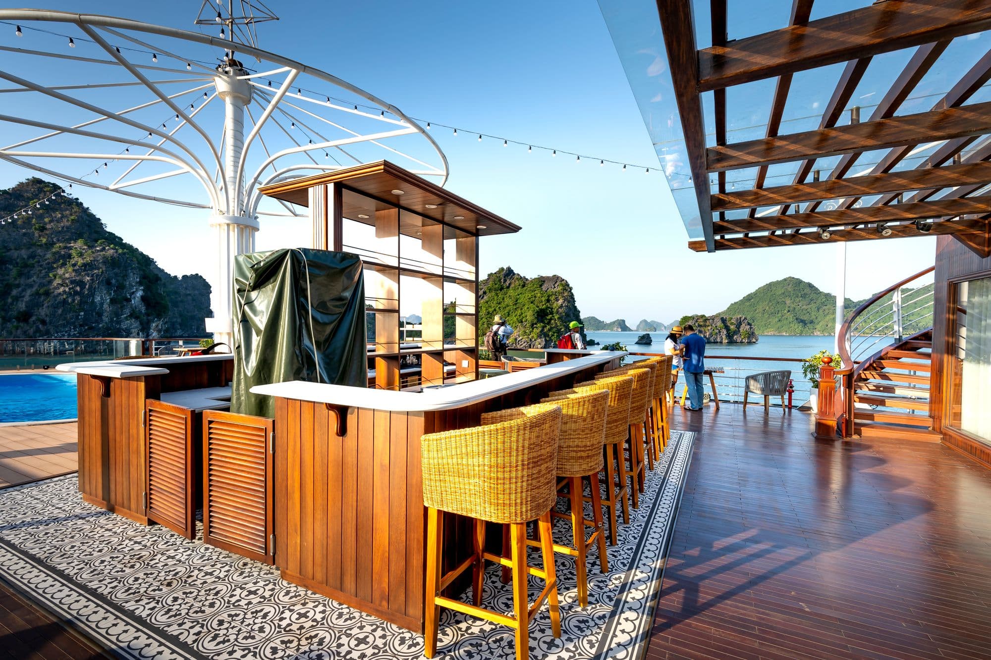 Wooden outdoor bar counter with louvered cabinet doors, a white bar top and wicker bar stools on a deck, with a fitted green cover over an appliance in the middle
