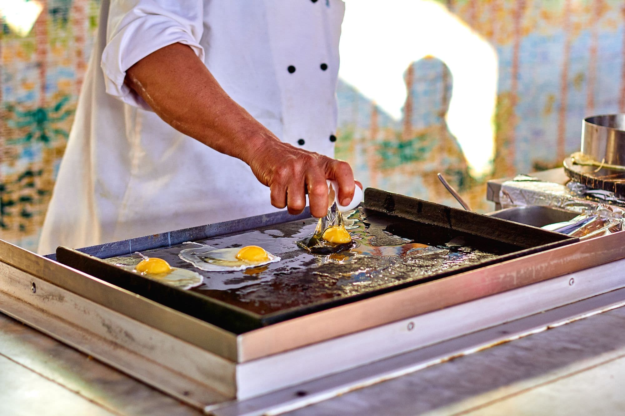 Cook in a white jacket cracking eggs onto an outdoor stainless flat-top griddle