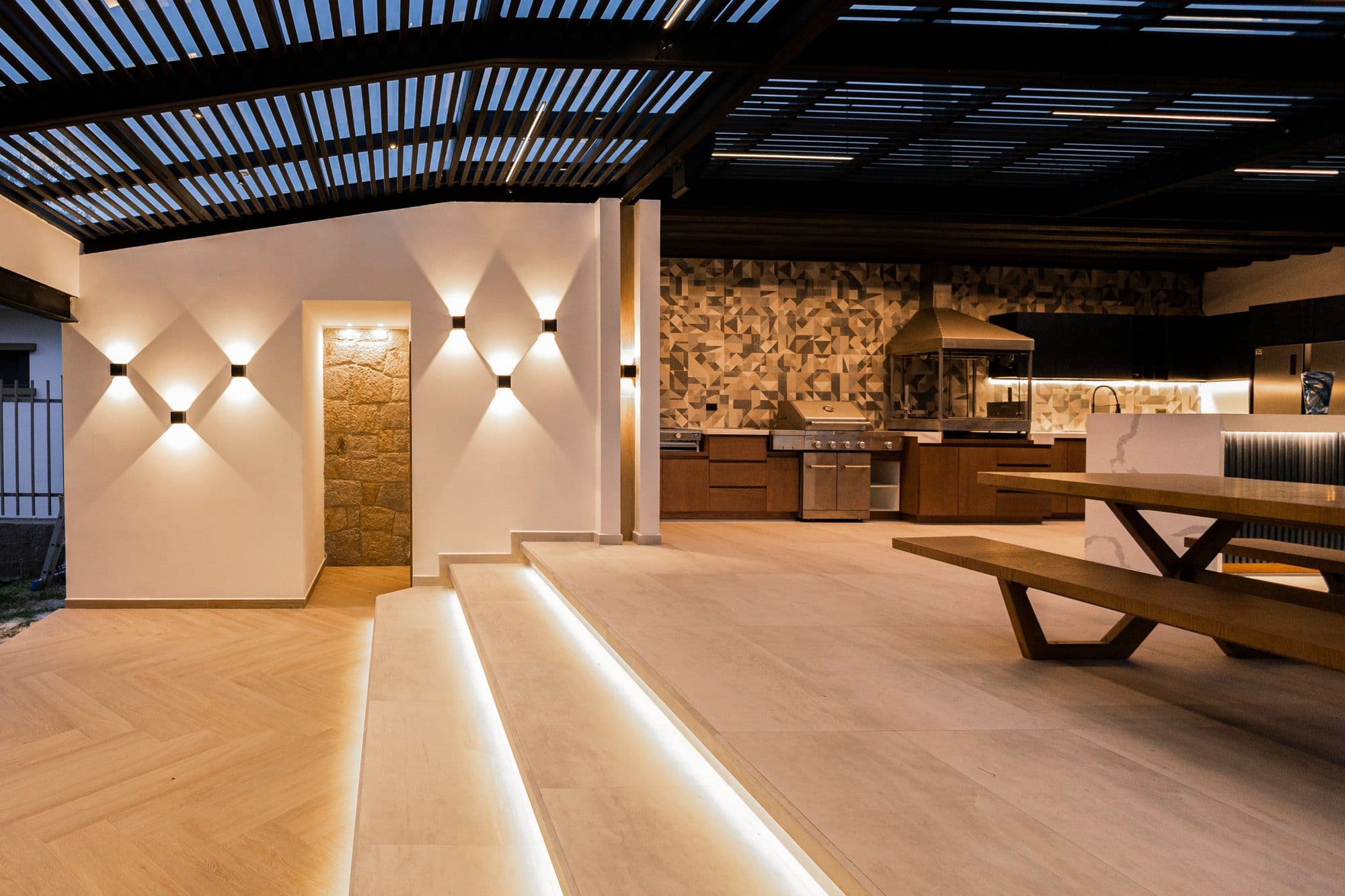 Louvered-roof outdoor kitchen pavilion lit by wall sconces and LED step lighting, with the grill run and tile backsplash in the background