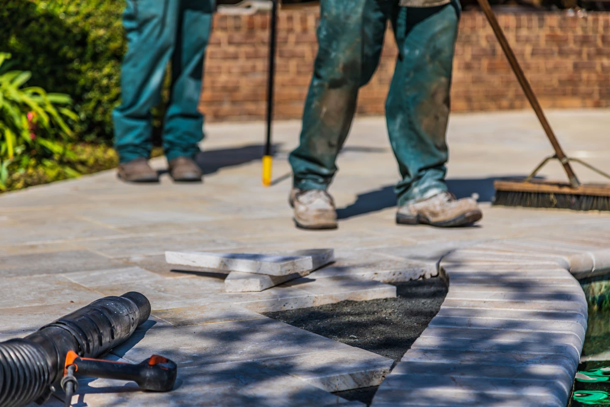 Workers laying and sweeping light stone pavers on a new patio, with a plate tool in the foreground
