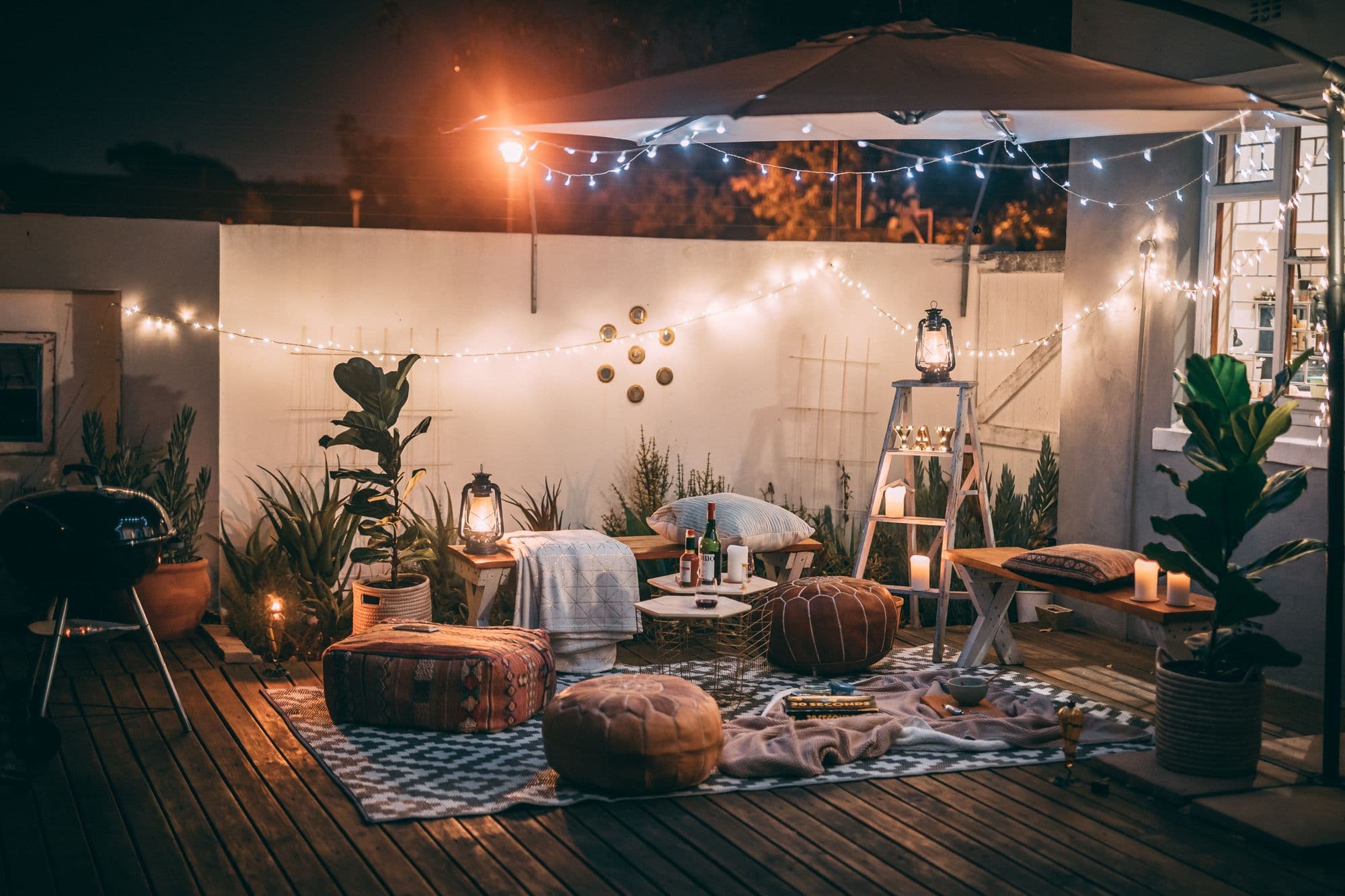 Small backyard deck at night lit by string lights and lanterns, with floor cushions, plants and a kettle grill on the left