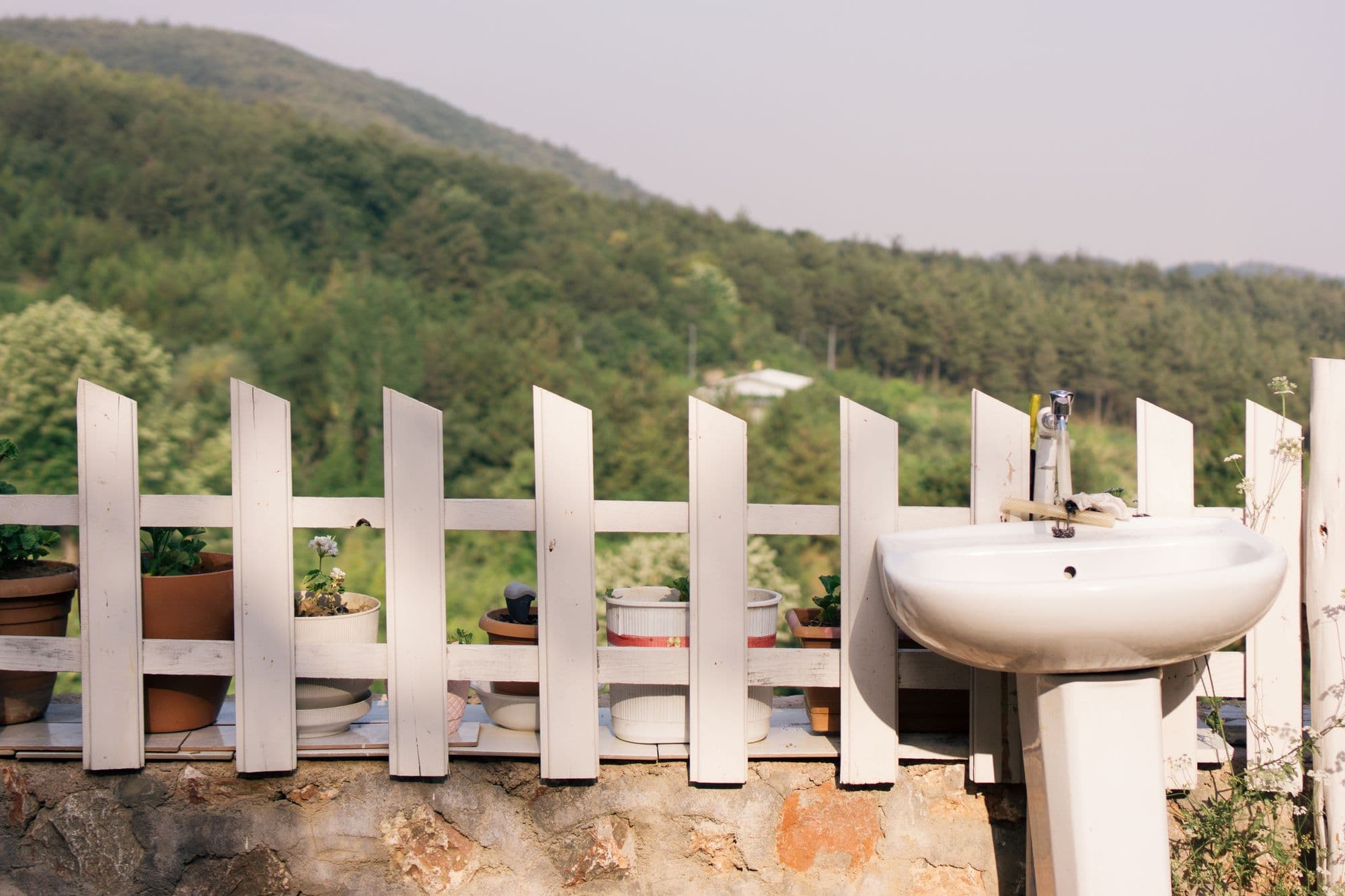 White pedestal sink set against a picket fence with potted plants and a forested hillside behind
