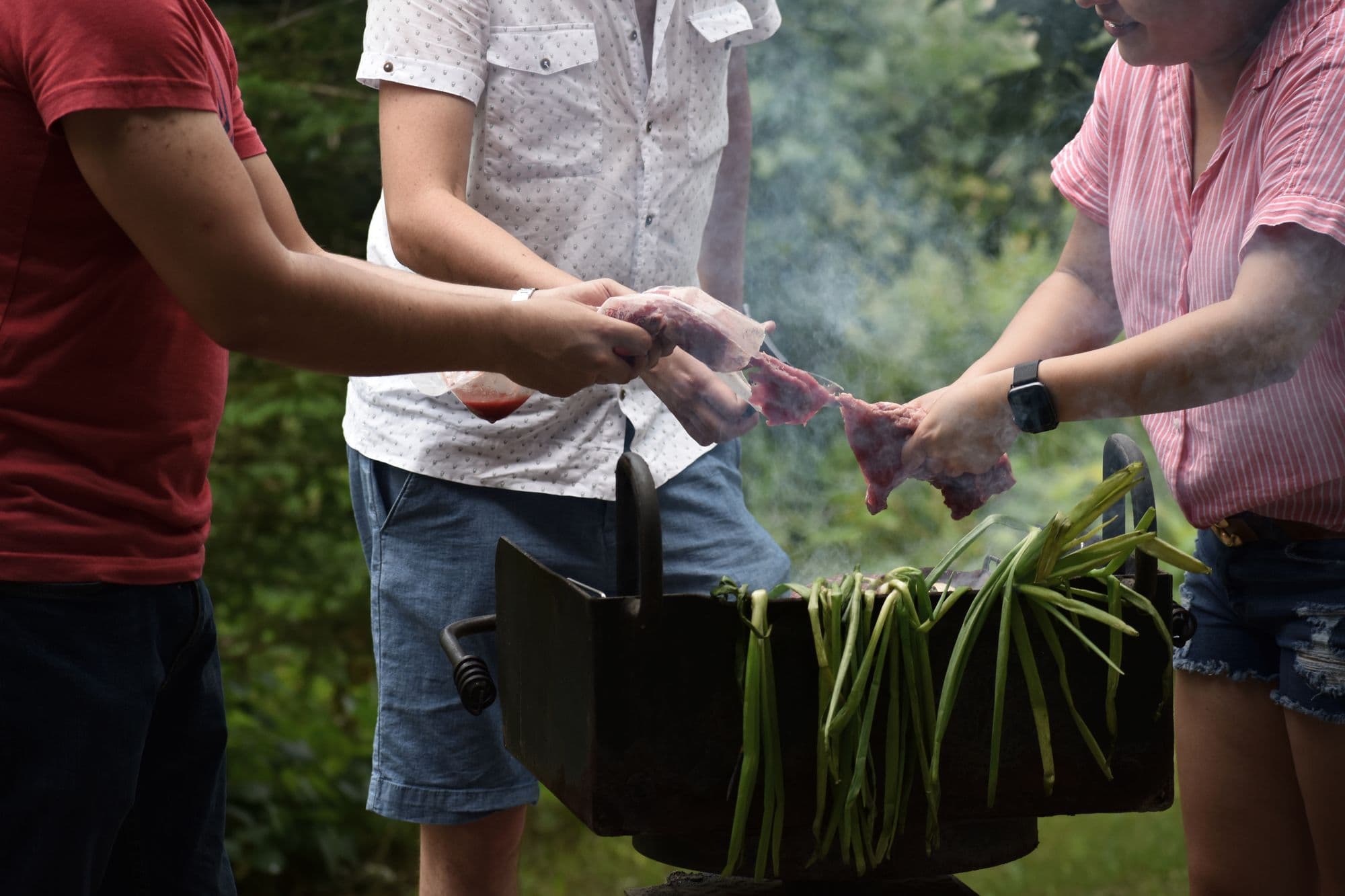 Three people putting meat and green onions on a smoking charcoal grill in a garden