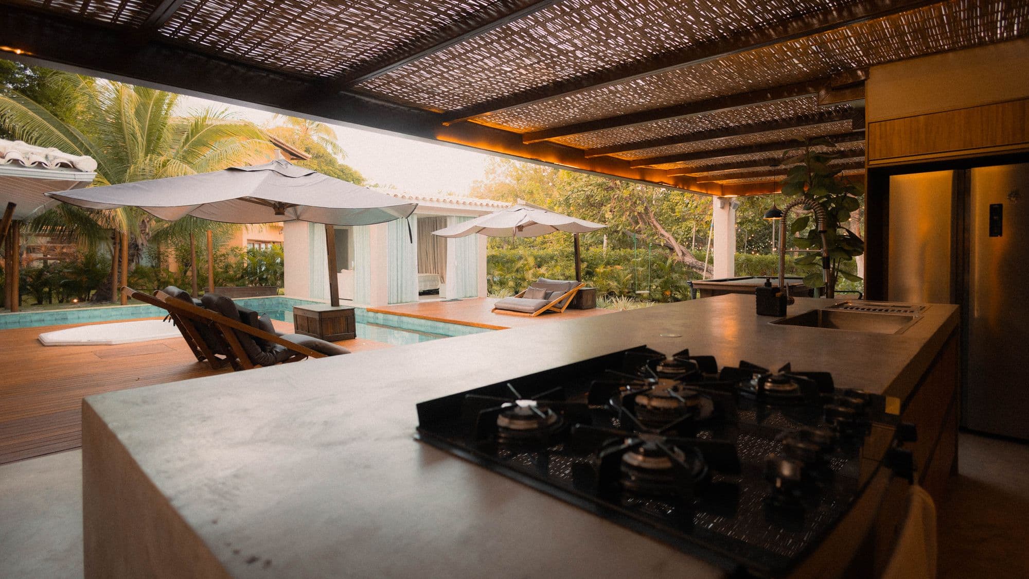 Concrete outdoor kitchen counter with a built-in gas cooktop under a slatted shade roof, looking out over a swimming pool and lounge chairs