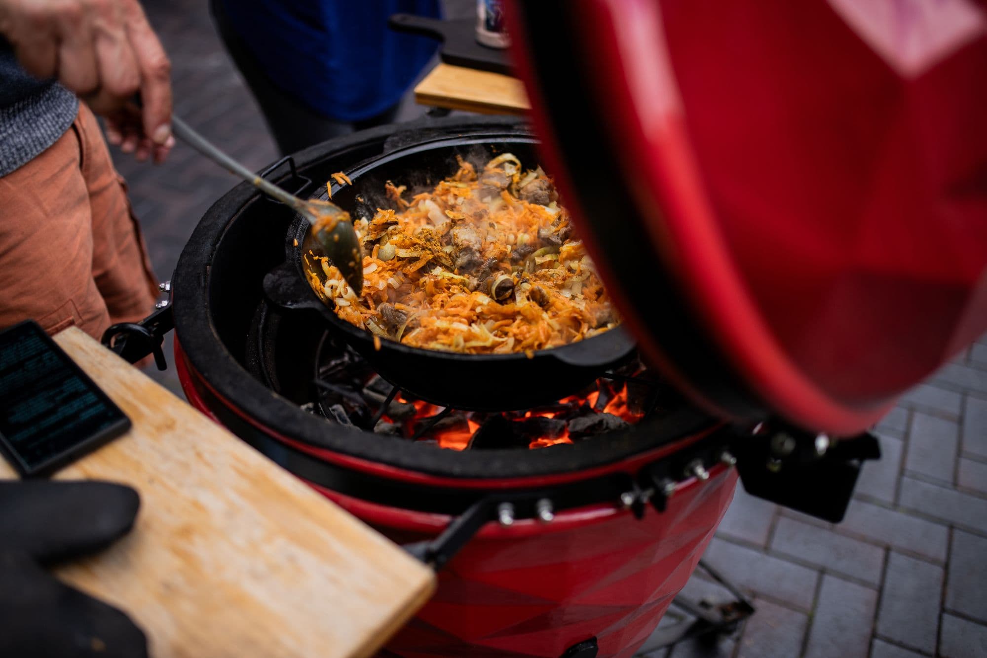 Red ceramic kamado grill with its lid open, a cast-iron pan of pilaf over glowing charcoal