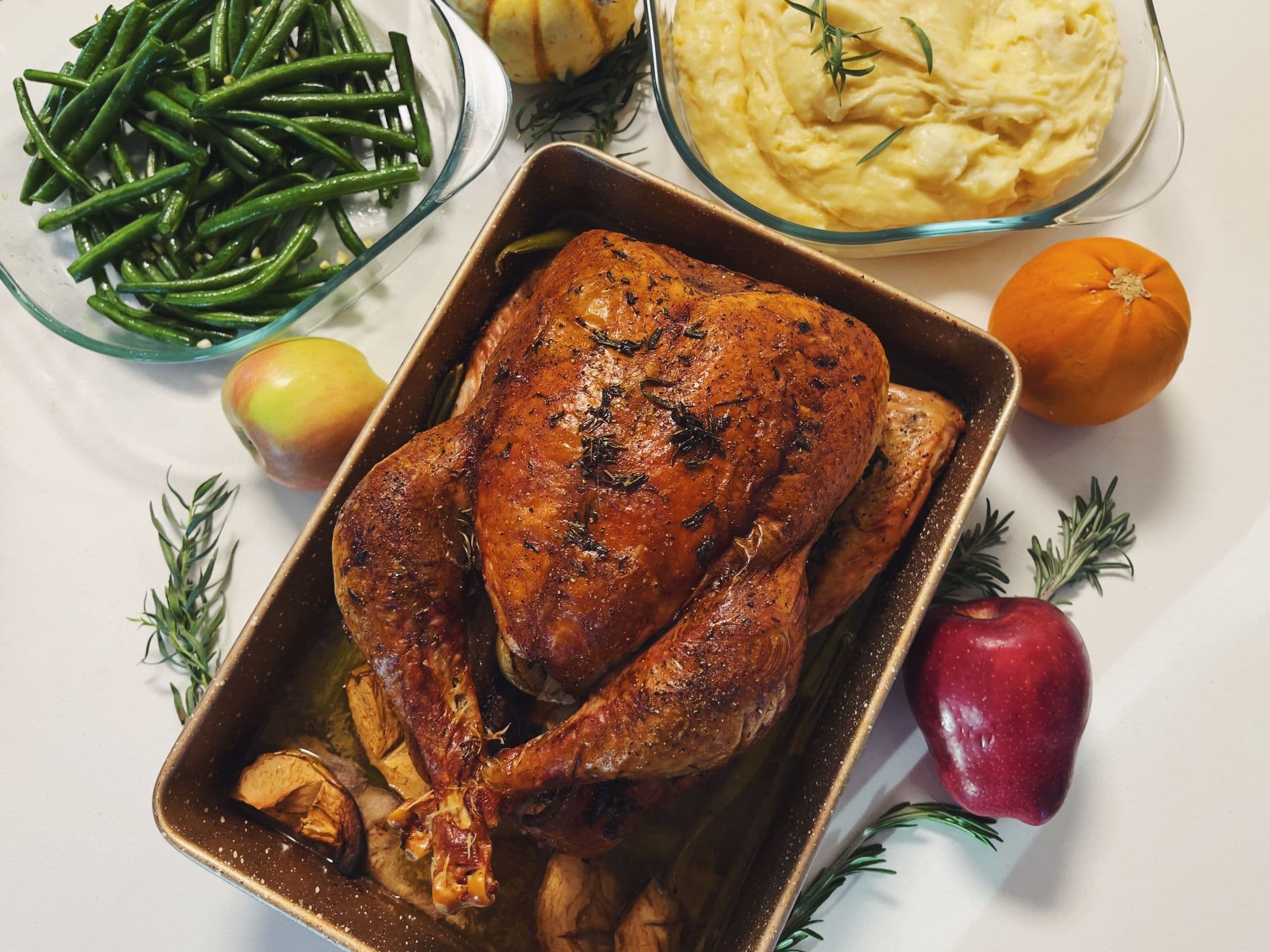 Roast turkey in a pan beside green beans, mashed potatoes, an orange and apples