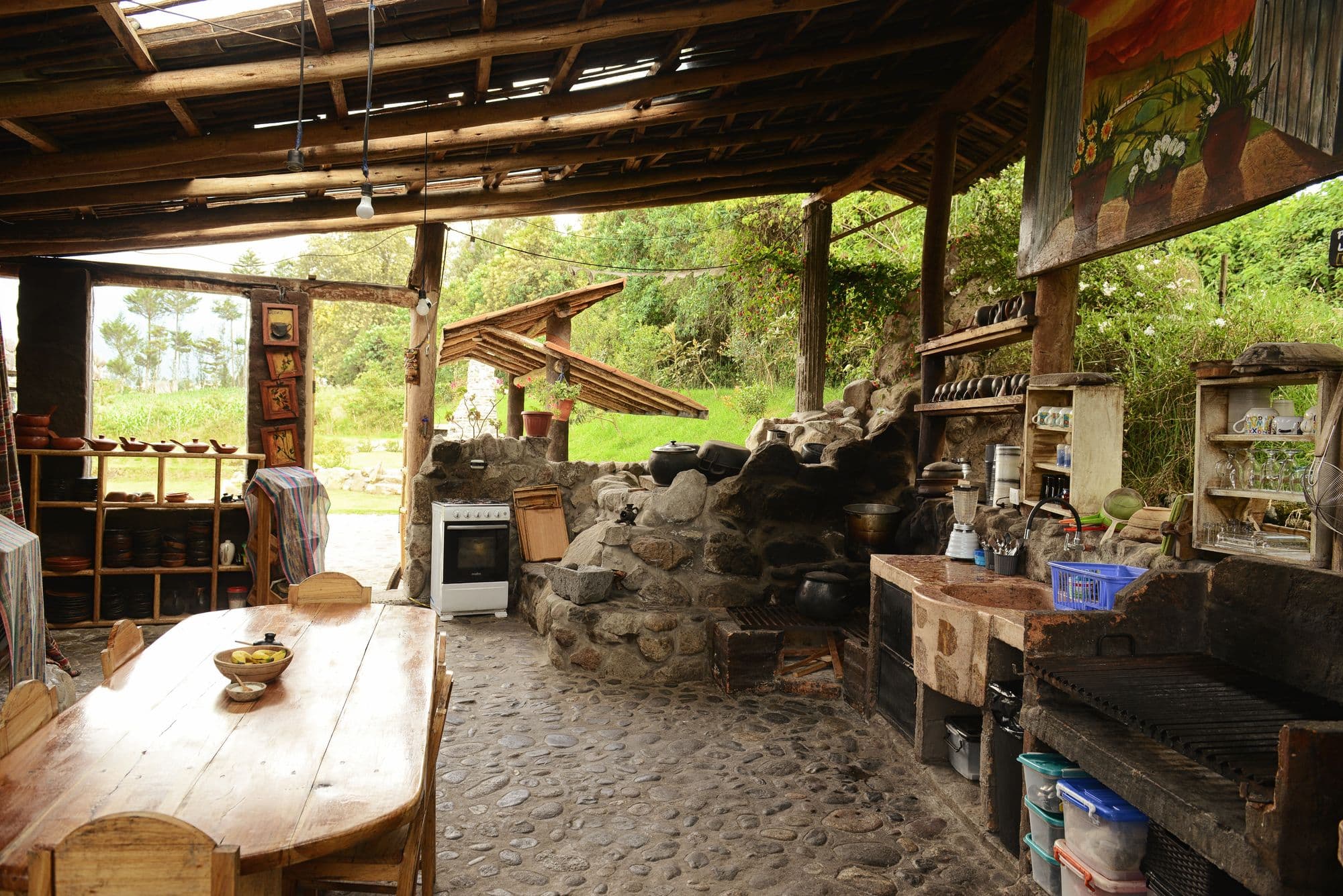 Large rustic covered outdoor kitchen with a fieldstone hearth, a charcoal grill, open shelving and a long wooden dining table on a cobblestone floor
