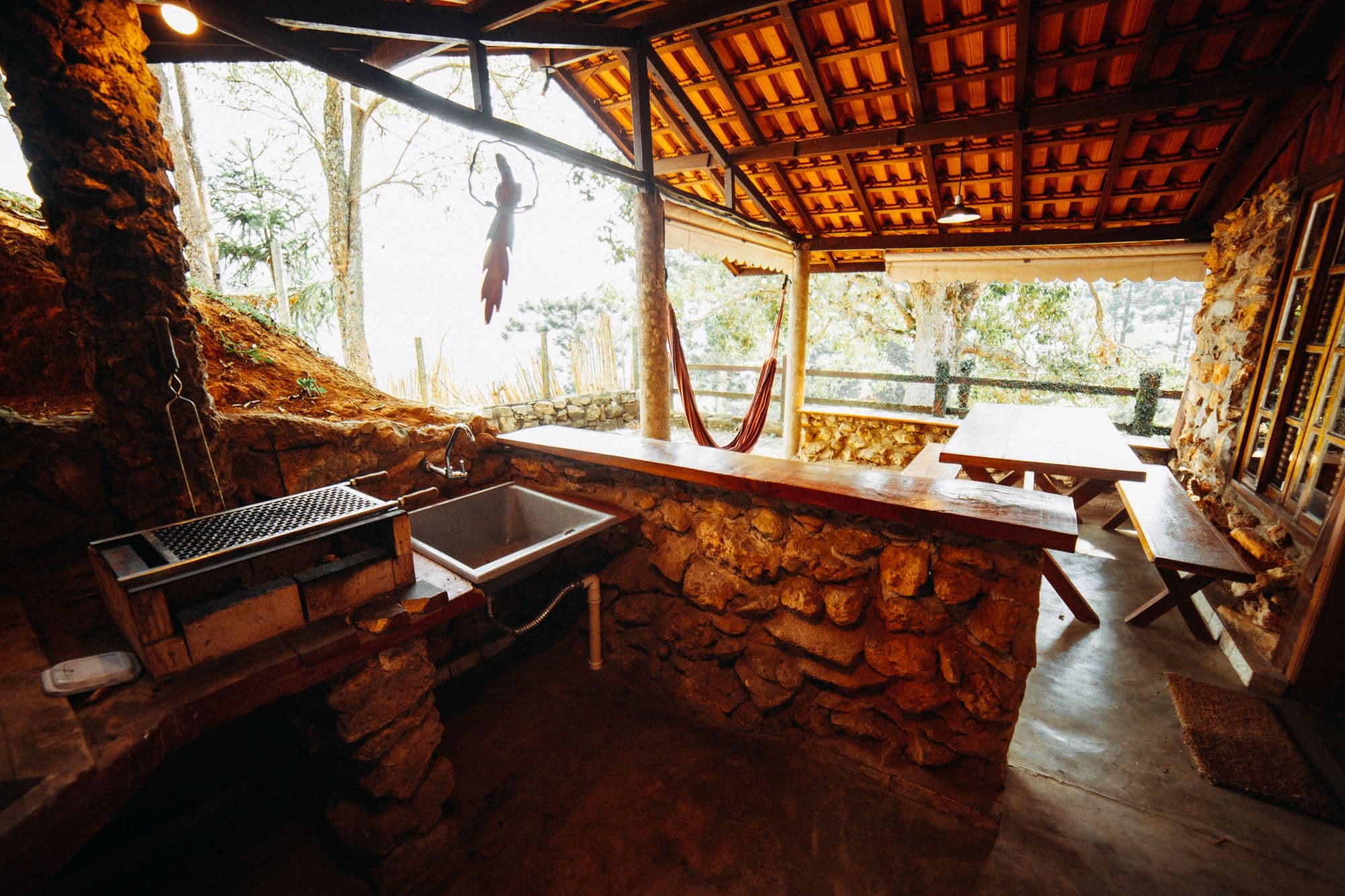 Rustic open-air kitchen with a stacked fieldstone counter, a charcoal grill grate, a stainless sink and a picnic table under a clay-tile roof