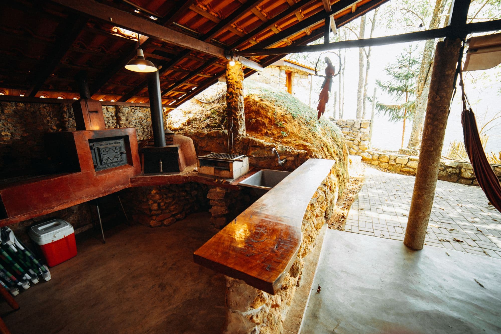 Rustic covered outdoor kitchen with a stone-and-clay wood-fired oven, a cast-iron stove, a stone sink counter and a polished wood bar top