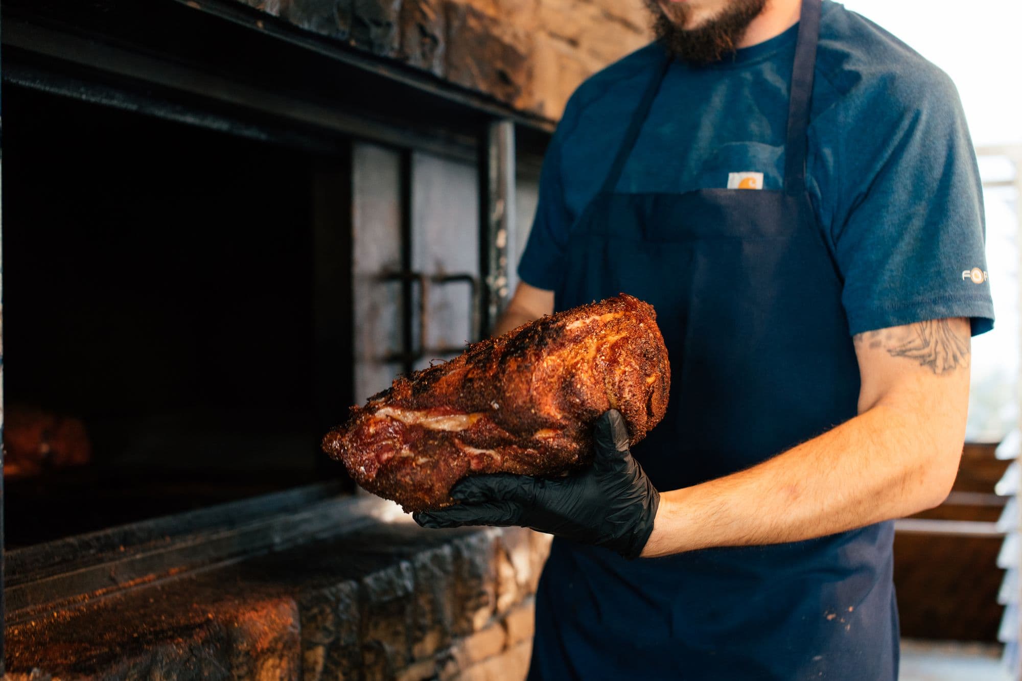 Gloved cook holding a bark-crusted smoked pork shoulder in front of a masonry barbecue pit
