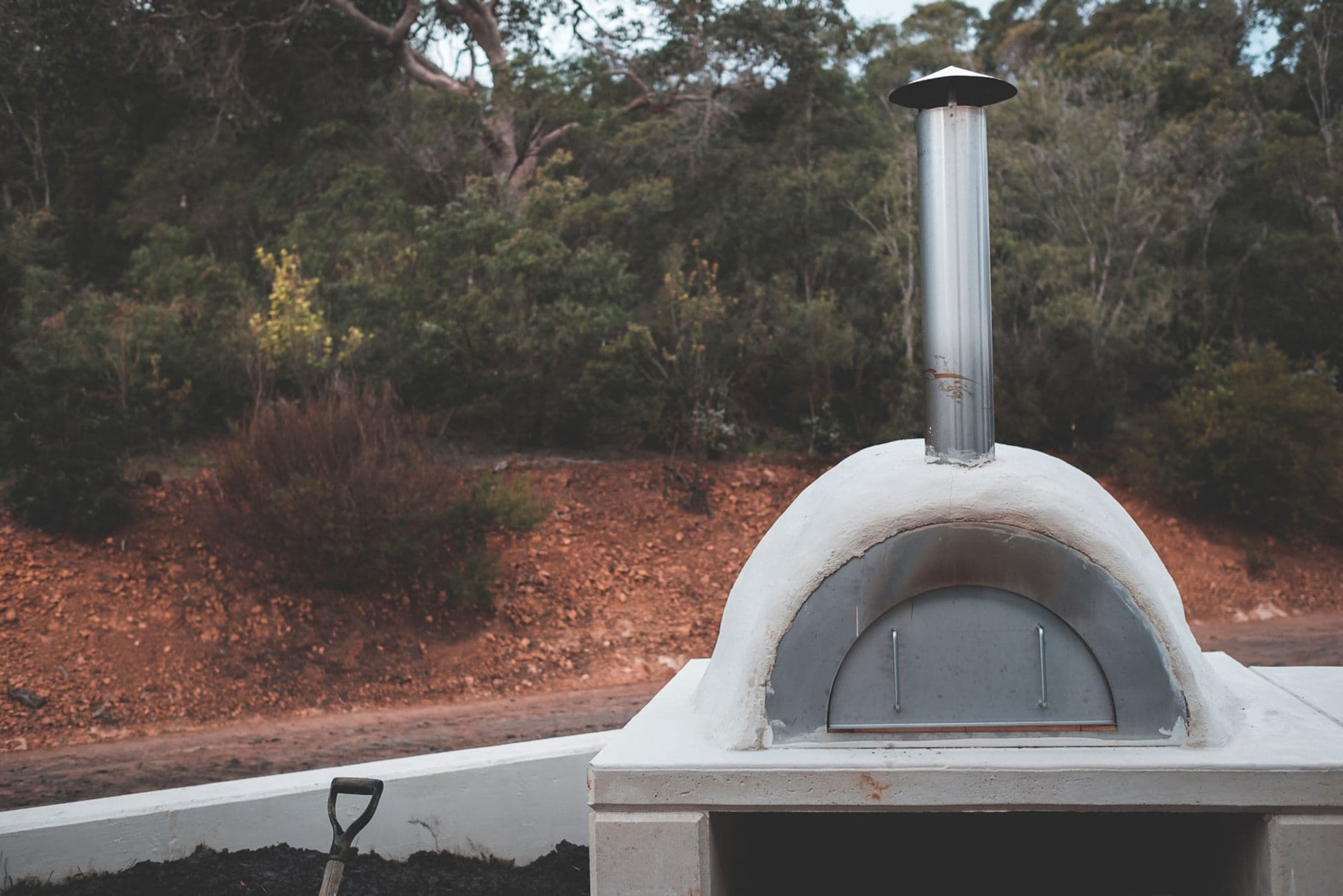 Dome pizza oven with a stainless door and tall flue on a white masonry base in a garden