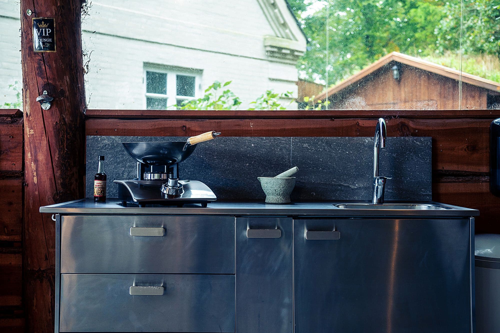 Compact stainless steel outdoor kitchen run with drawer and door fronts, a wok on a gas burner and an undermount sink with a tall faucet against a wooden fence