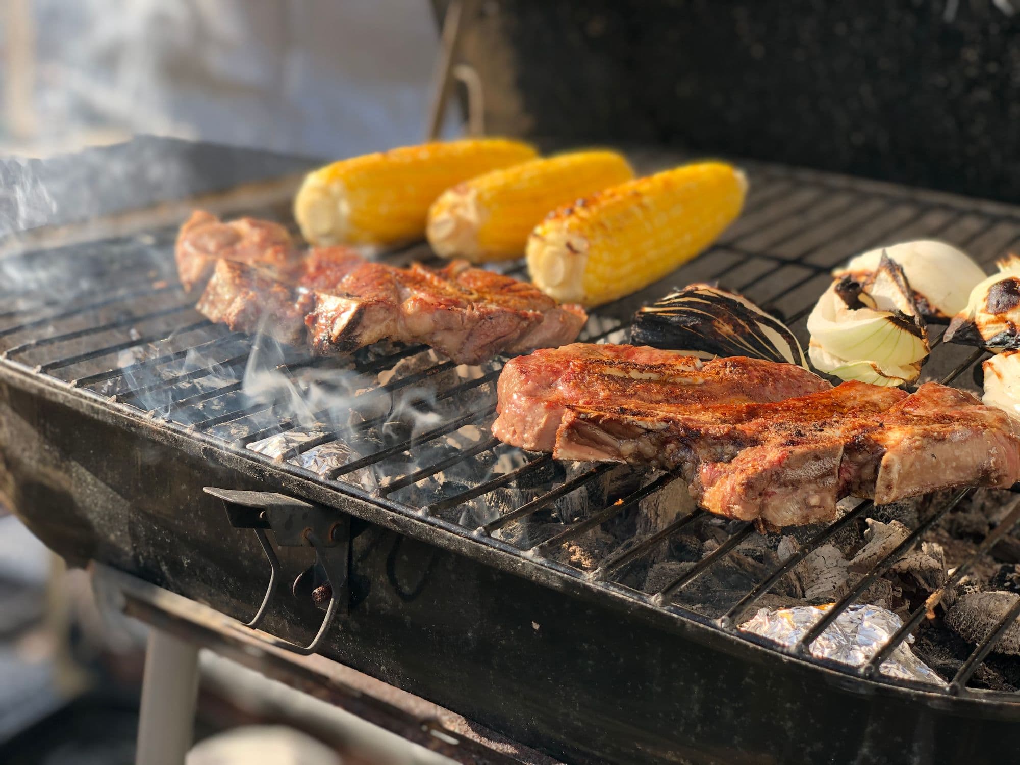 Steaks, corn on the cob and halved onions on a smoking grill grate