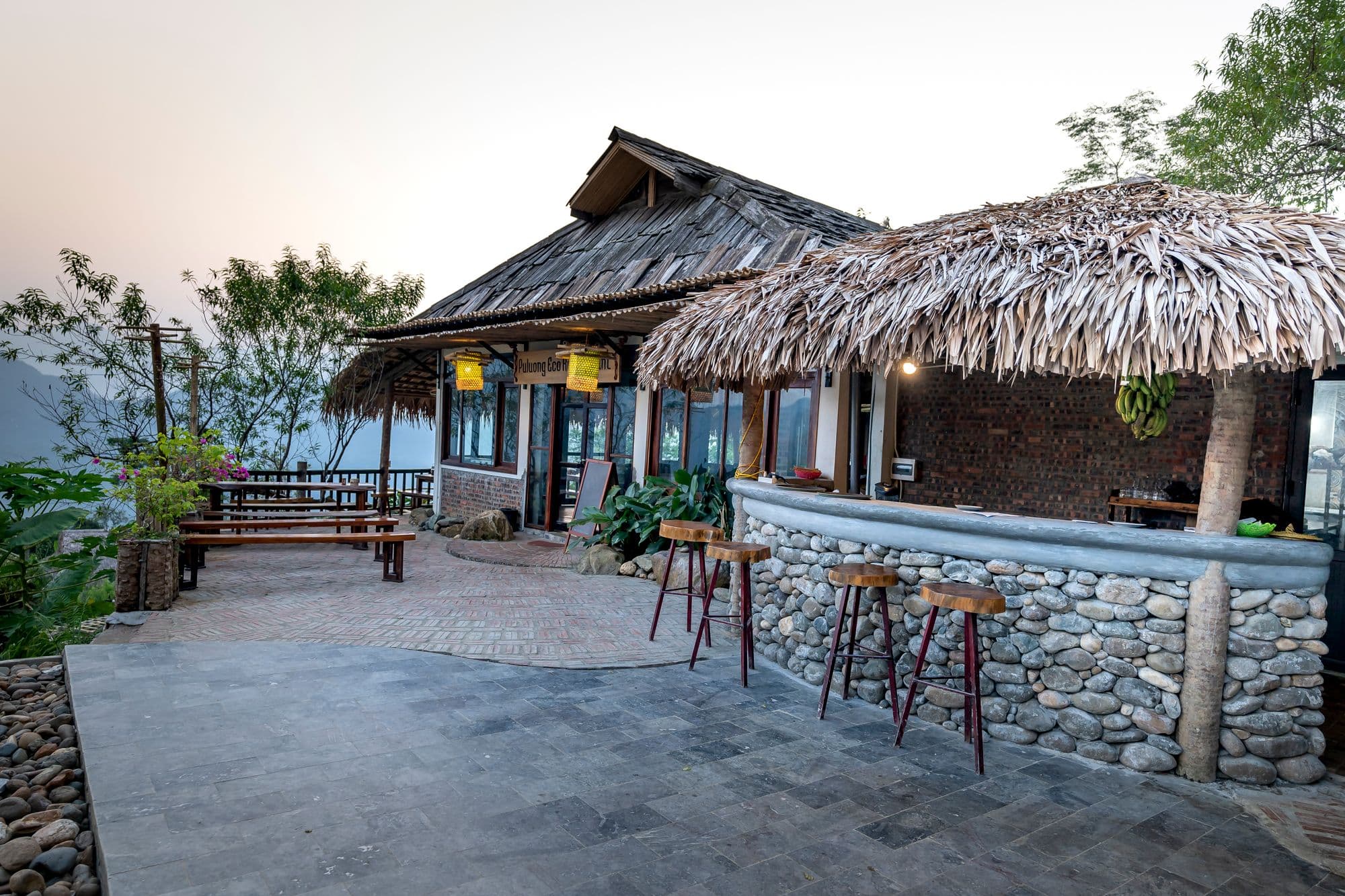 Curved river-stone bar counter with wooden stools in front of a thatched-roof building on a stone patio