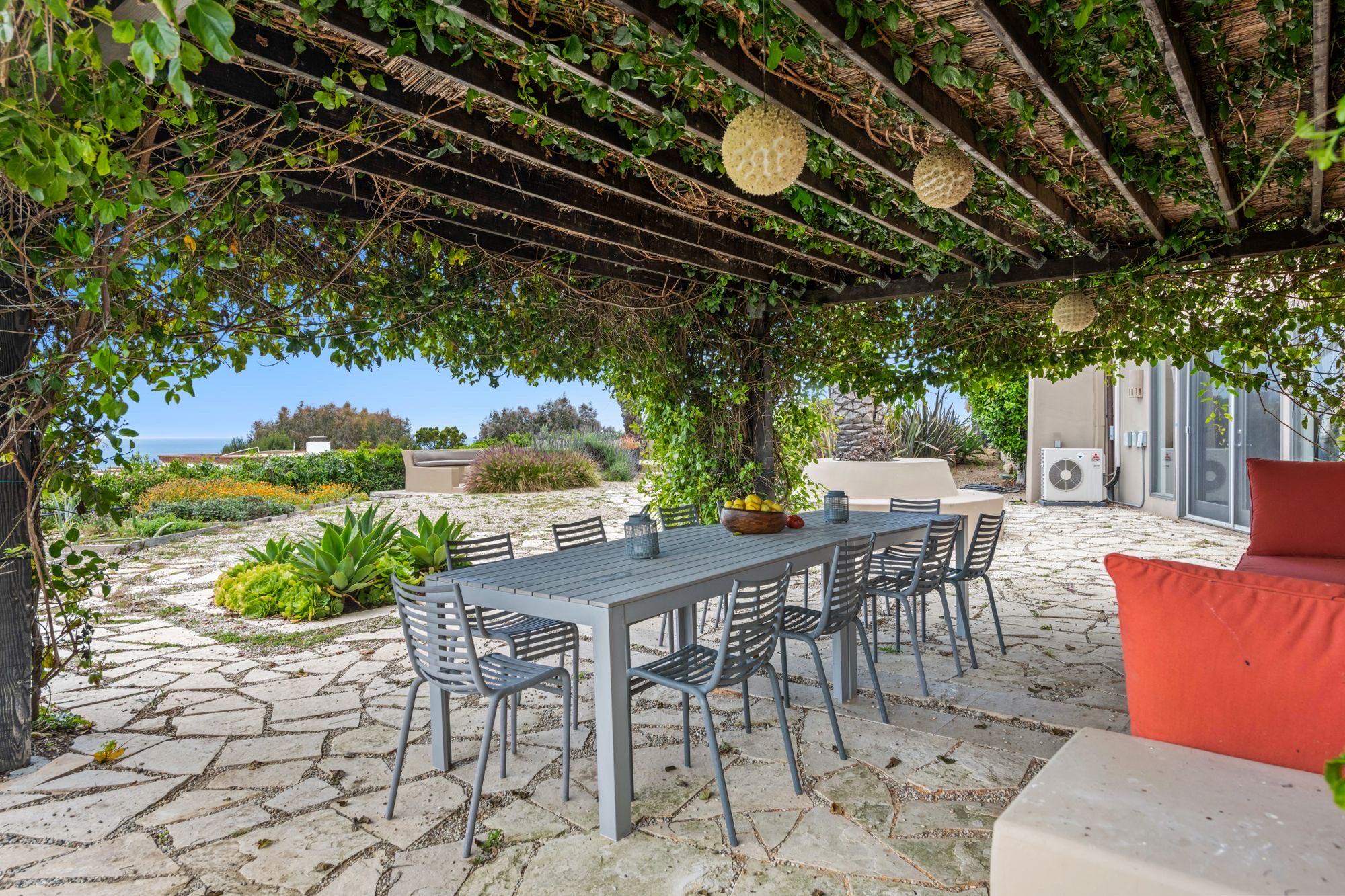 Outdoor dining table and chairs on a flagstone patio under a vine-covered pergola with hanging lanterns