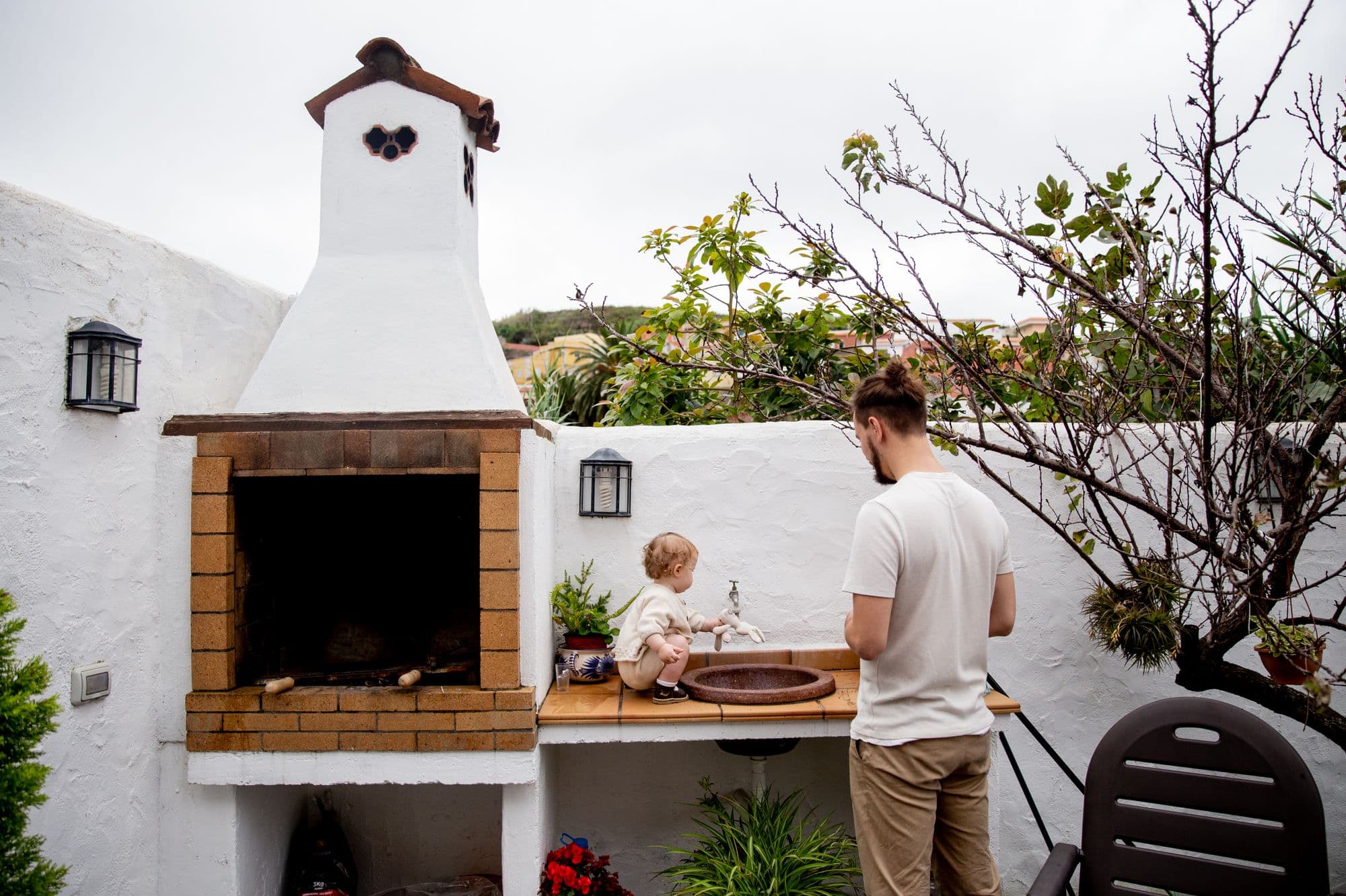 Whitewashed masonry courtyard barbecue with a chimney, a wood-topped counter and a copper sink, with a man and a toddler at the sink