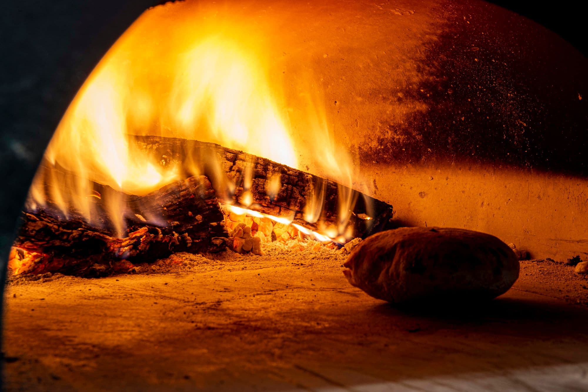 Close-up of logs burning inside a domed wood-fired oven with a bread loaf on the hearth