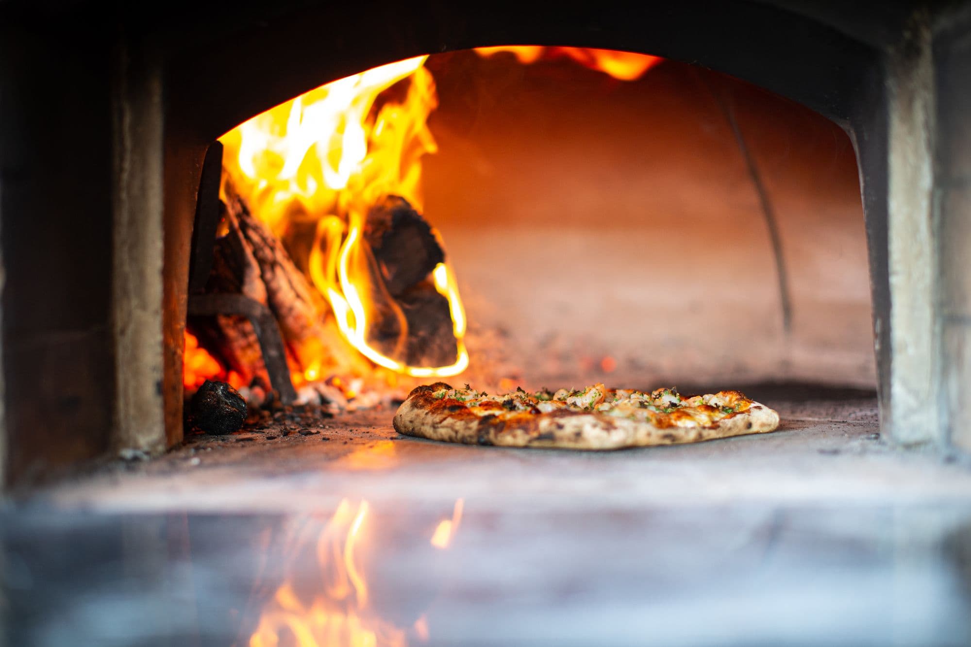 Pizza baking on the deck of a wood-fired oven beside burning logs